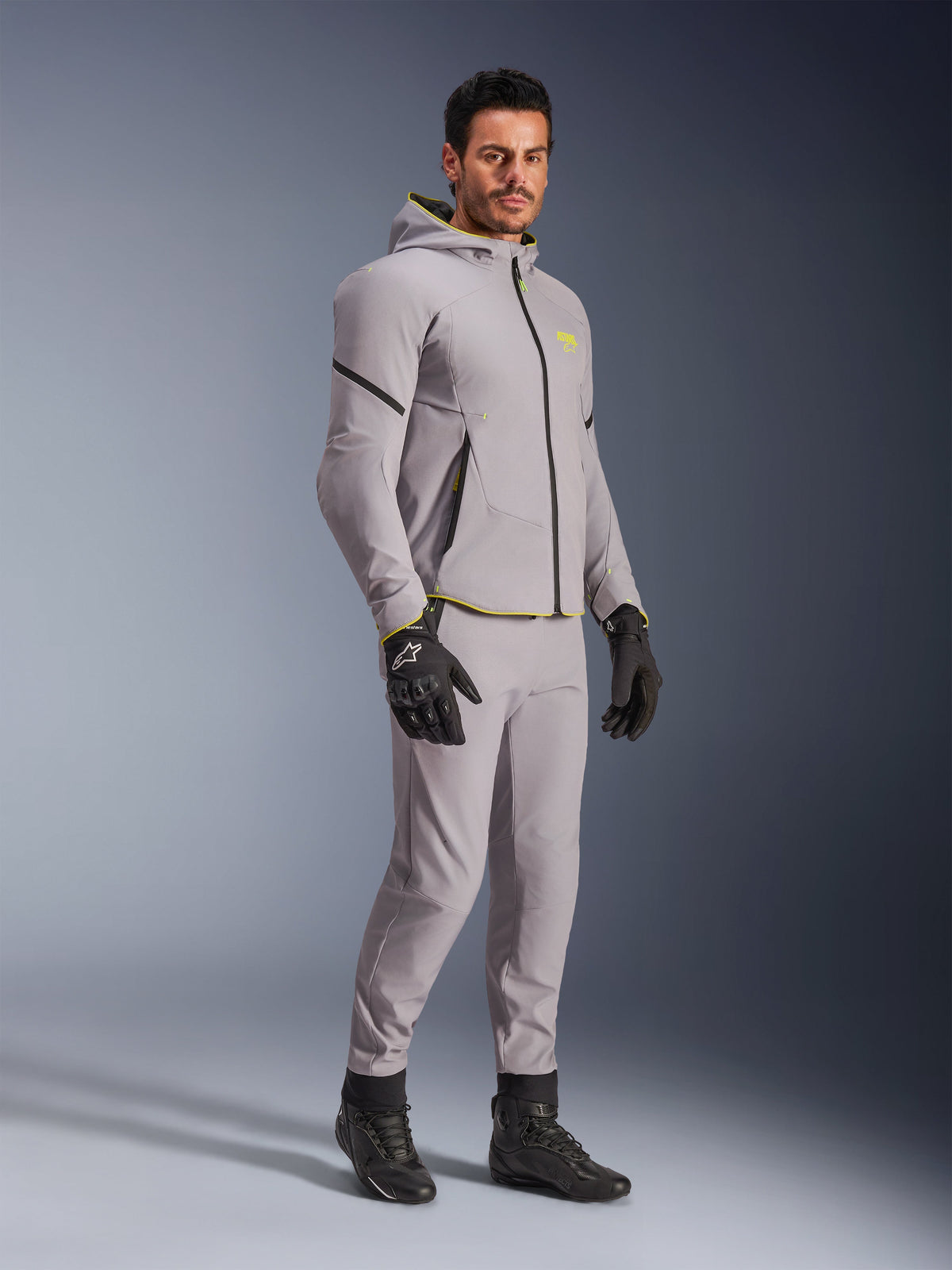 Chaqueta impermeable Alpinestars Aeroshell, gris medio y amarillo flúor, usada por un modelo con pantalones grises a juego, guantes de moto negros y botas negras, equipamiento de moto moderno estilo sudadera con capucha para conducción casual y deportiva.