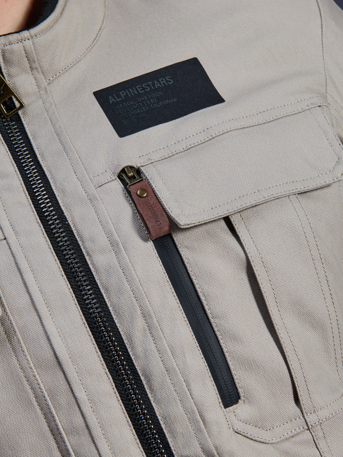 Alpinestars Borrego Drystar® Jacket, Warm Gray, close-up detail of chest area featuring a black Alpinestars branding patch, chest pocket with flap, and a vertical zipper with a brown leather pull tab, durable gray textile canvas construction.