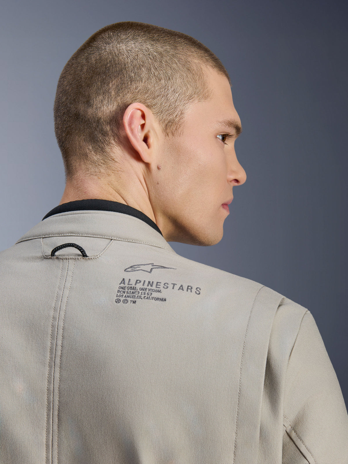 Alpinestars Borrego Drystar® Jacket in Warm Gray, close-up of back shoulder, textured waxed-canvas material, featuring black Alpinestars logo and heritage text, worn by model, adventure motorcycle jacket