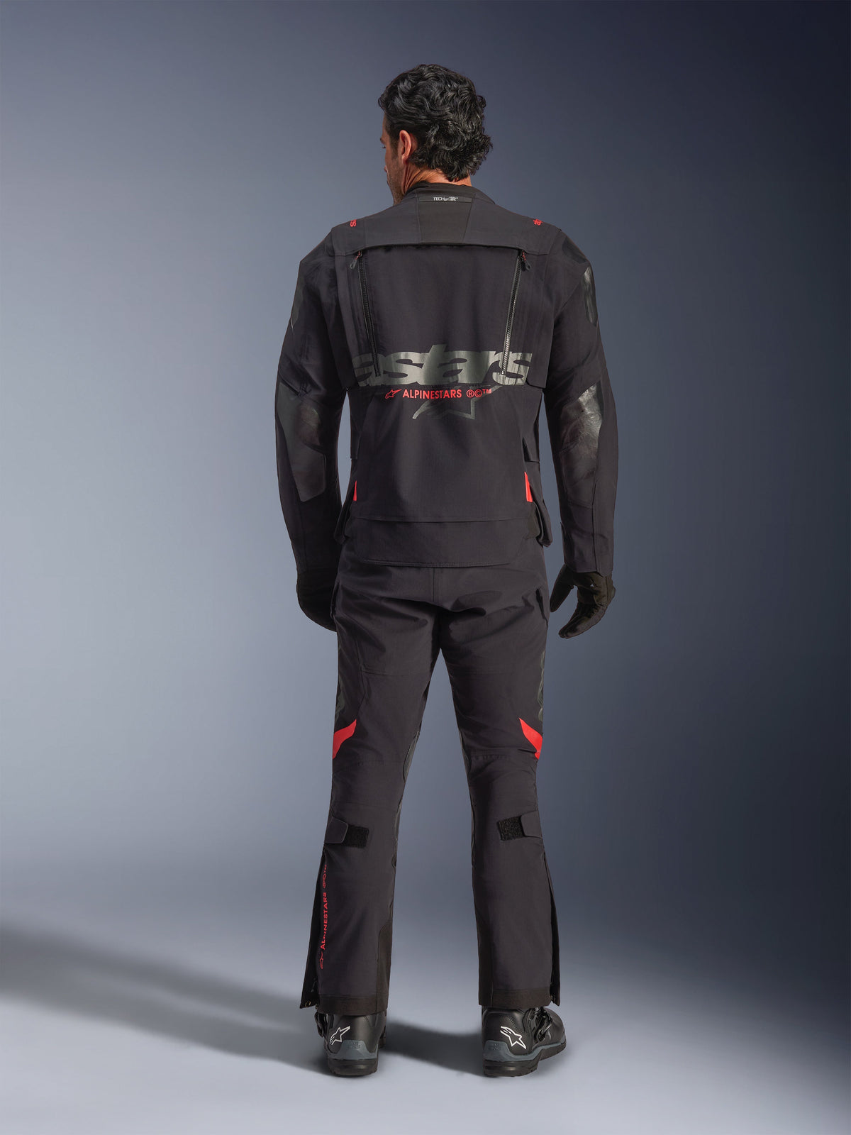 Un piloto visto desde atrás vistiendo la chaqueta laminada Alpinestars Halo Pro Drystar® XF en Black Dark Shadow, con un gran logotipo astars gris y marca Alpinestars en rojo, paneles de ventilación modulares en la espalda, combinada con pantalones de aventura a juego y botas negras.