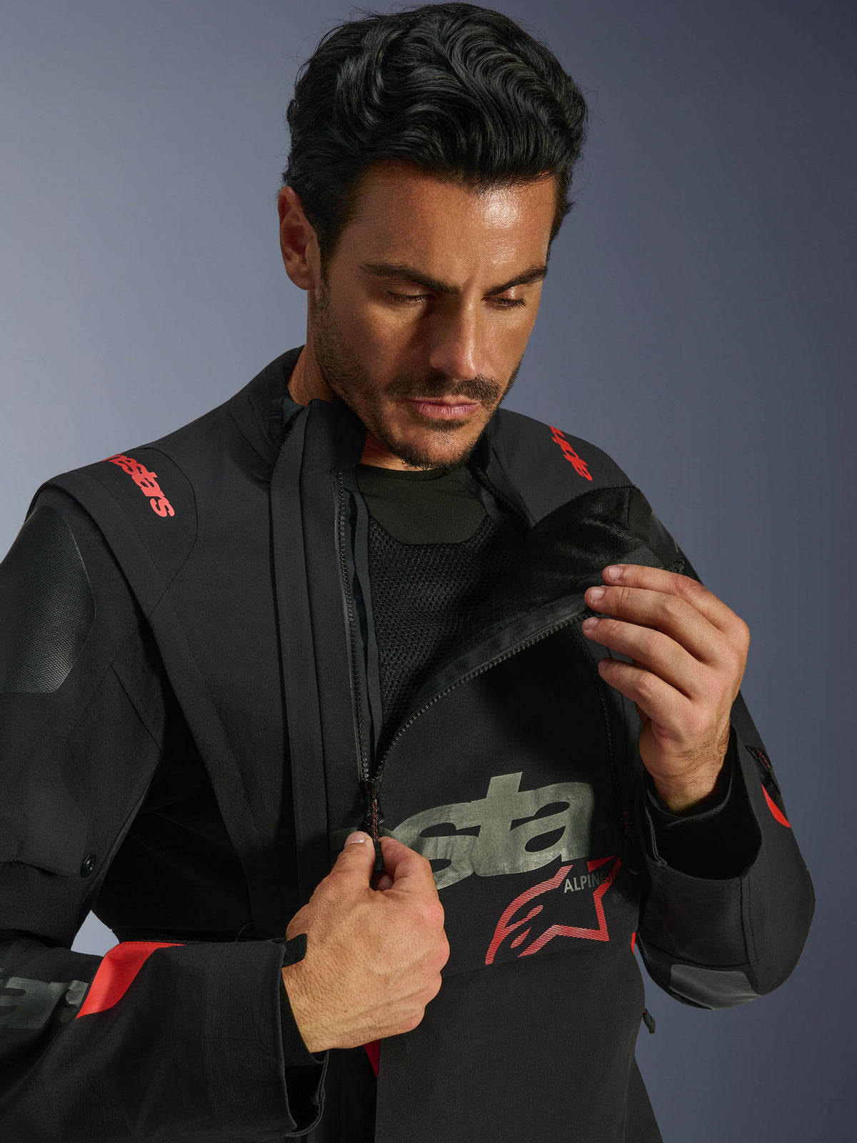 Chaqueta laminada Alpinestars Halo Pro Drystar® XF, Black Dark Shadow, modelo demostrando el sistema de ventilación modular al abrir la cremallera del panel frontal asimétrico del pecho, chaqueta de aventura con marca Alpinestars en rojo y detalles de malla técnica.