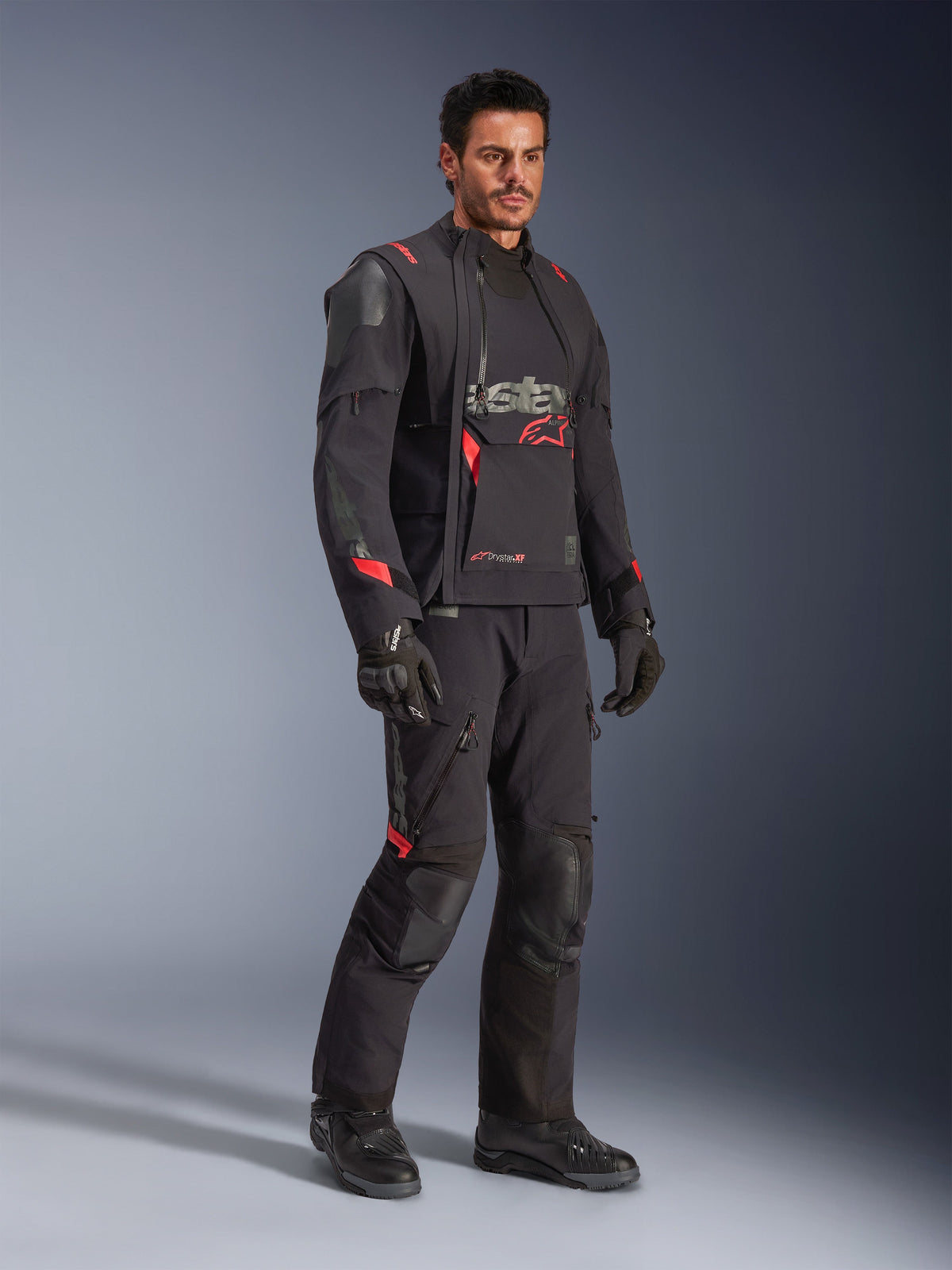 Piloto con la chaqueta laminada Alpinestars Halo Pro Drystar® XF en Black Dark Shadow, equipamiento de aventura completo con pantalones, guantes y botas a juego, paneles frontales asimétricos modulares y detalles rojos de la marca.