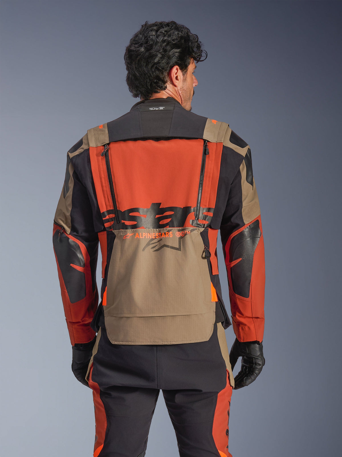 Chaqueta laminada Alpinestars Halo Pro Drystar® XF, combinación de colores Walnut Rust Black, vista trasera, con un gran panel trasero naranja con la marca ASTARS en negro, bolsillo de almacenamiento inferior color nogal y cuello preparado para Tech-Air®, usada con pantalones de aventura a juego y guantes negros.