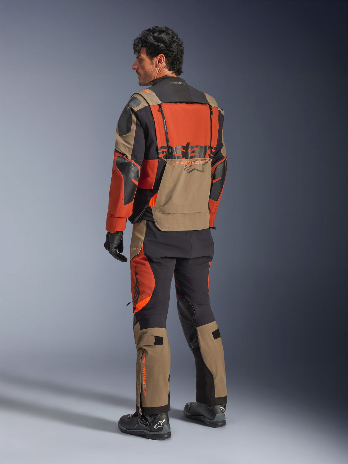 Chaqueta laminada Alpinestars Halo Pro Drystar® XF, chaqueta de moto de aventura en Walnut Rust Black, vista trasera usada con pantalones a juego y botas negras, con material elástico técnico 3L, paneles de ventilación modulares y logotipos naranjas en la parte posterior