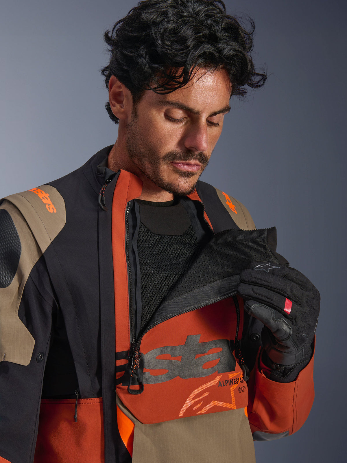 Chaqueta laminada Alpinestars Halo Pro Drystar® XF, combinación de colores Walnut Rust Black, modelo masculino demostrando el sistema de ventilación modular al abrir el panel frontal del pecho para revelar la malla negra, chaqueta técnica de moto de aventura con detalles en óxido y tostado.