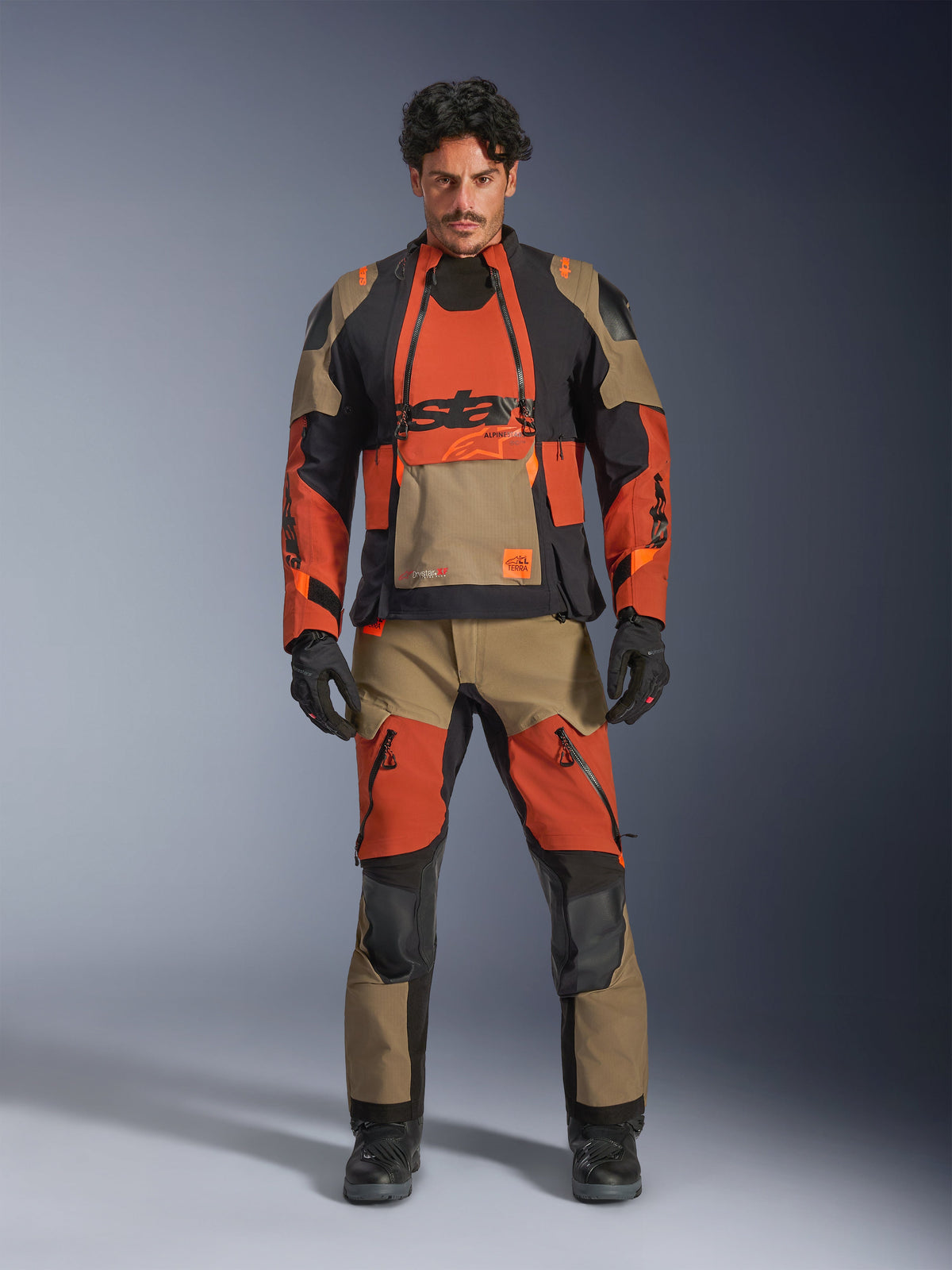 Modelo masculino vistiendo la chaqueta laminada Alpinestars Halo Pro Drystar® XF y pantalones a juego en Walnut Rust Black, equipamiento modular de aventura para motociclismo con paneles de ventilación frontales y cremalleras, combinado con guantes y botas de moto negros.