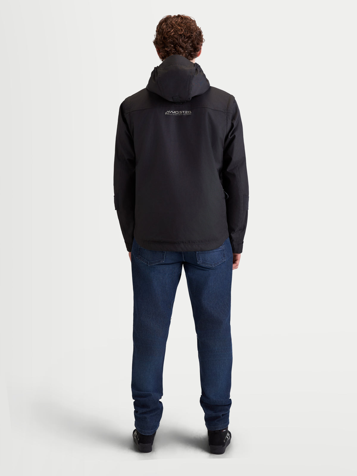 Alpinestars Morfex Parka Waterproof Jacket, chaqueta de moto urbana contemporánea para hombre en color negro, vista trasera en un modelo que lleva la chaqueta con capucha integrada y vaqueros de moto casuales