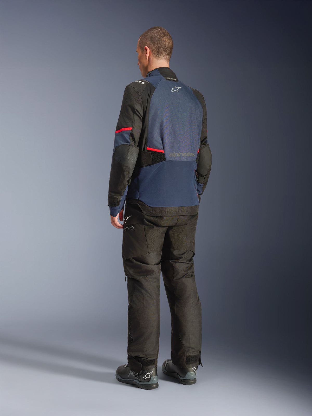 Chaqueta Alpinestars Andes V4 Drystar®, azul noche y azul índigo con paneles negros y detalles rojos, vista trasera en tres cuartos, combinada con pantalones de aventura y botas negras a juego, construcción en tejido técnico con logotipo de Alpinestars en la parte superior trasera y dobladillo posterior alargado para mayor cobertura.