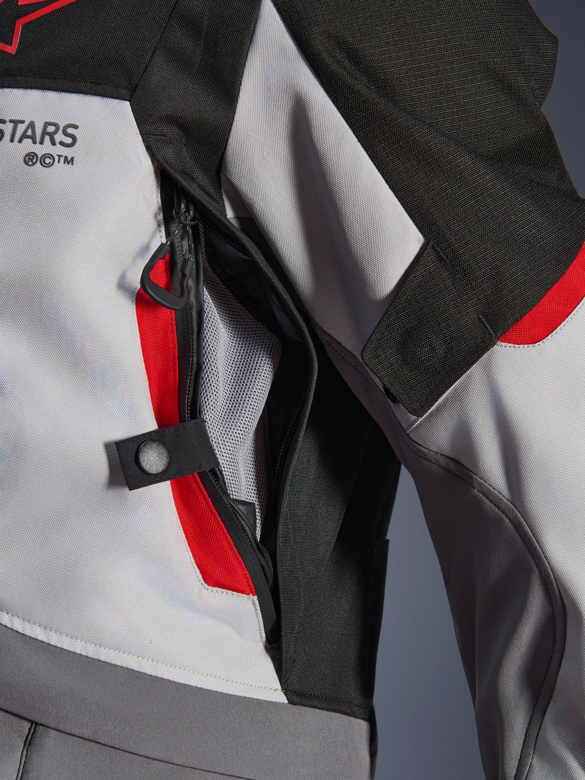 Detalle de primer plano de la chaqueta Alpinestars Andes V4 Drystar® en gris oscuro, gris hielo y rojo brillante, que destaca una abertura de ventilación con cremallera en el pecho con forro de malla y una correa de ajuste con botón a presión.