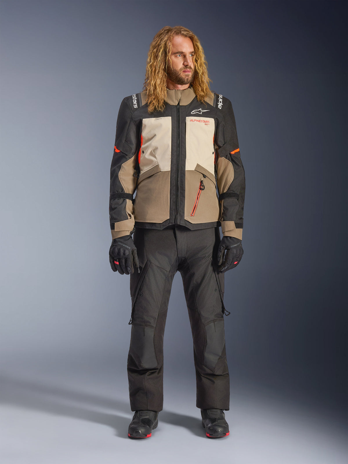 Chaqueta Alpinestars Andes V4 Drystar®, Walnut Mastic Black, combinada con pantalones y guantes de motociclismo negros, con un diseño de aventura de múltiples paneles, detalles en naranja y el logotipo de Alpinestars, equipamiento textil técnico para conducir en todo tipo de clima.