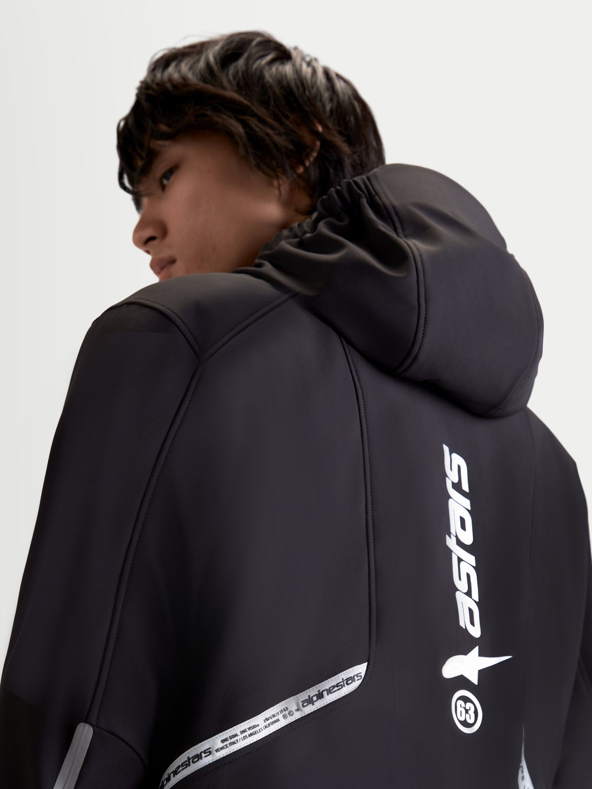 Alpinestars C 5 Drystar chaqueta, chaqueta de moto urbana contemporánea para hombre en color negro, vista trasera de cerca que muestra la capucha integrada y detalles de marca reflectantes usada por un modelo