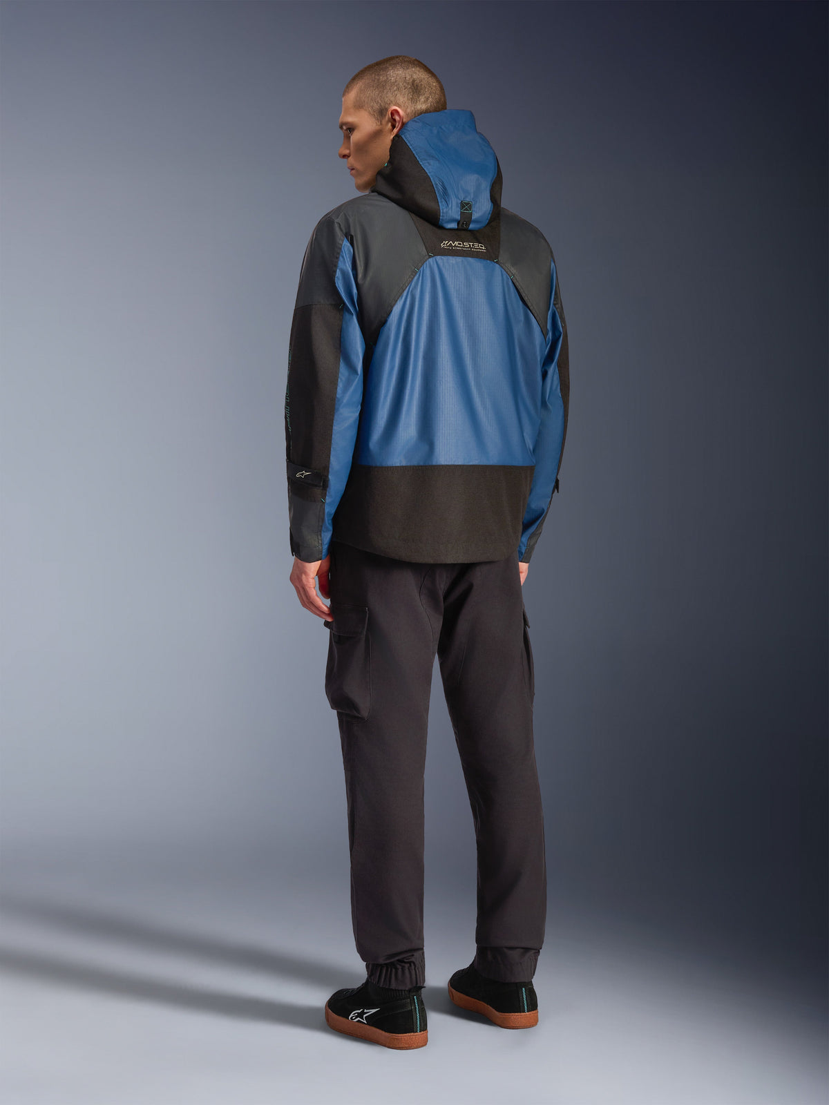 Vista posterior de un modelo masculino que viste la chaqueta impermeable Alpinestars Mospher en color Black y Dark Petrol, con un diseño con capucha y confección táctica en ripstop, combinada con pantalones cargo negros y zapatillas.