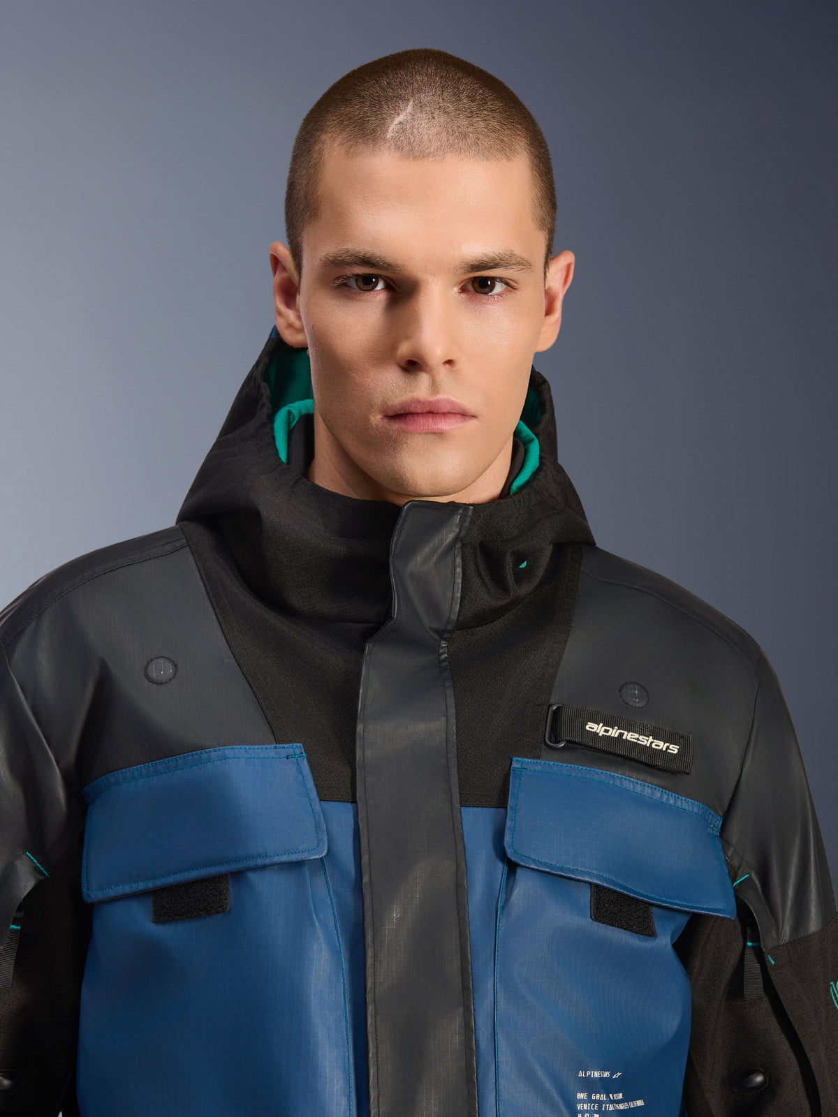 Chaqueta impermeable Alpinestars Mospher, negro y petróleo oscuro, primer plano de modelo masculino vistiendo la parka de estilo utilitario, con bolsillos en el pecho azul oscuro, capucha negra con forro verde azulado y logotipo de Alpinestars en el pecho.