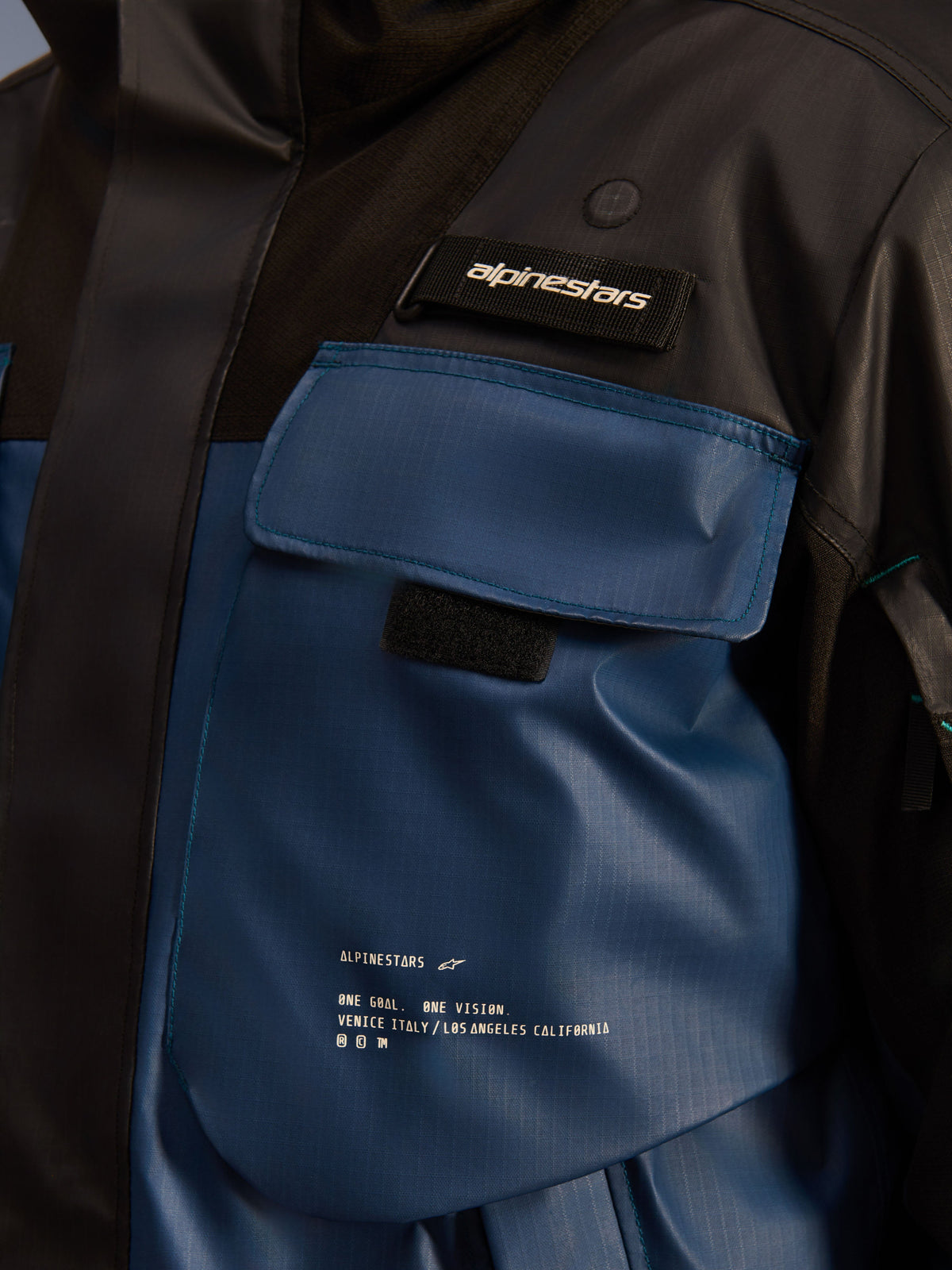 Chaqueta Alpinestars Mospher Waterproof, detalle en primer plano de la zona del bolsillo del pecho, negro y Dark Petrol, tejido ripstop duradero con marca 'One Goal. One Vision.' impresa, diseño funcional con bolsillos y pestaña con el logotipo de Alpinestars