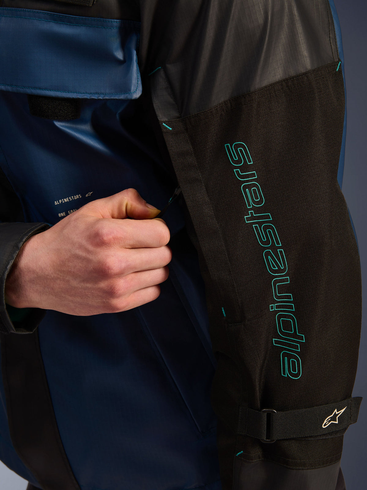 Chaqueta impermeable Alpinestars Mospher en color Black Dark Petrol, detalle en primer plano de la manga con el logotipo de Alpinestars en turquesa, una mano ajustando la cremallera de ventilación, con tejido ripstop duradero y una correa ajustable para el brazo