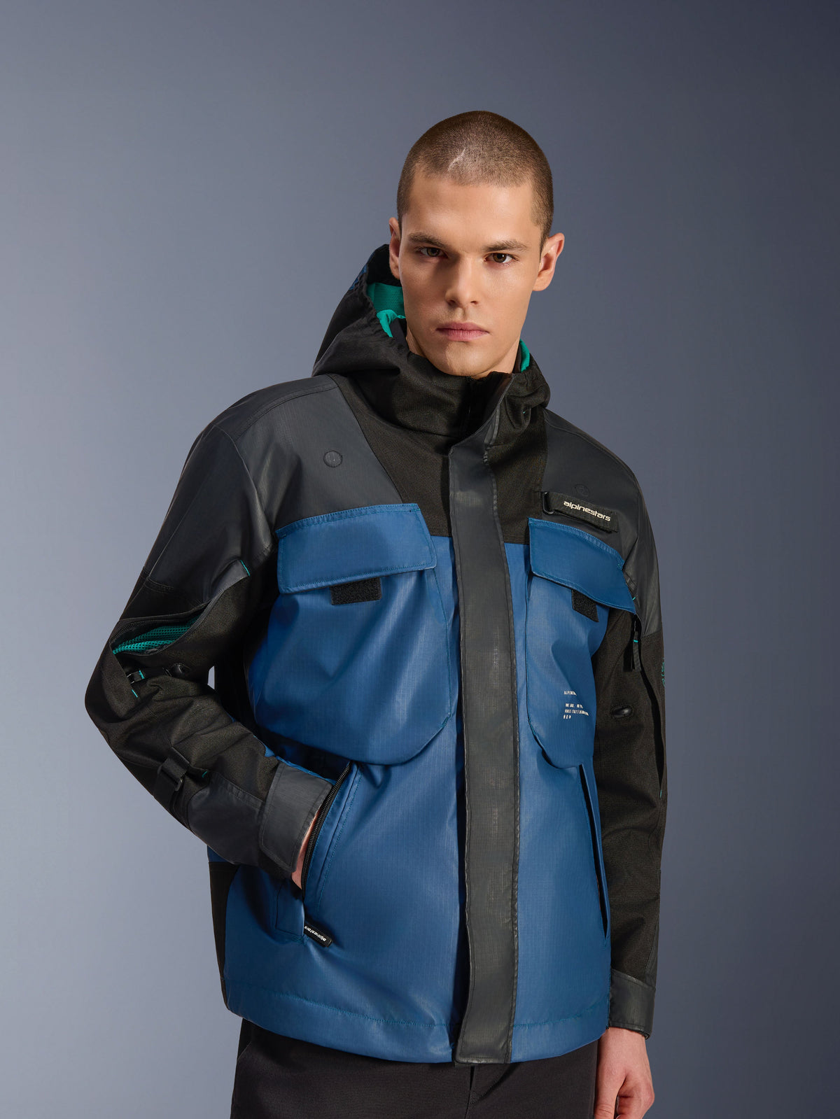 Modelo masculino con la chaqueta impermeable Alpinestars Mospher en negro y Dark Petrol, con grandes bolsillos utilitarios en el pecho, diseño con capucha y forro verde azulado, y tejido técnico ripstop para desplazamientos urbanos en moto