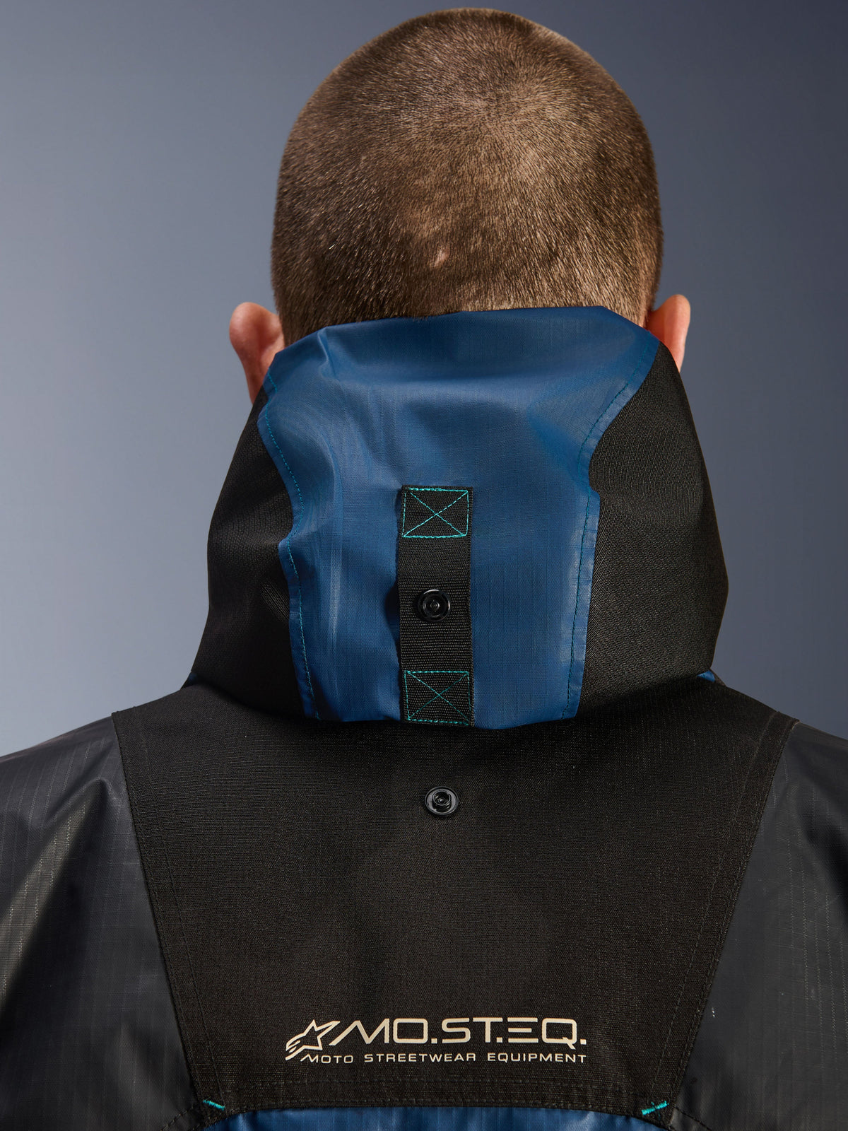 Chaqueta impermeable Alpinestars Mospher, detalle de la vista trasera que muestra el sistema de anclaje de la capucha, color Negro y Azul Petróleo Oscuro, con el logotipo de MO.ST.EQ. Moto Streetwear Equipment, material textil ripstop duradero, equipamiento técnico para motociclistas urbanos