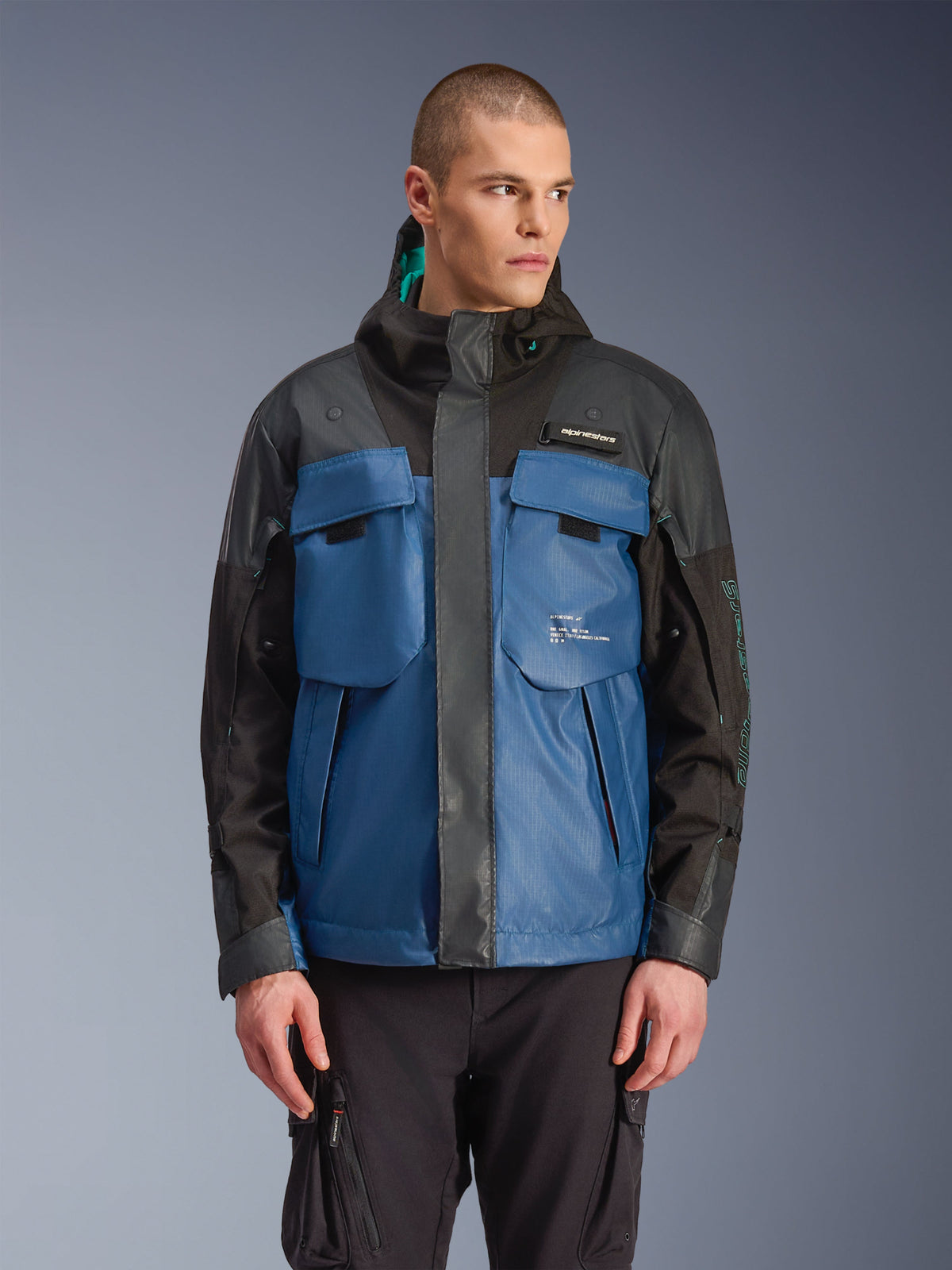 Alpinestars Mospher Waterproof Jacket, parka urbana para moto, negro y azul petrol oscuro, con grandes bolsillos tácticos en el pecho, cuello alto con capucha, textura técnica ripstop, vista frontal en modelo