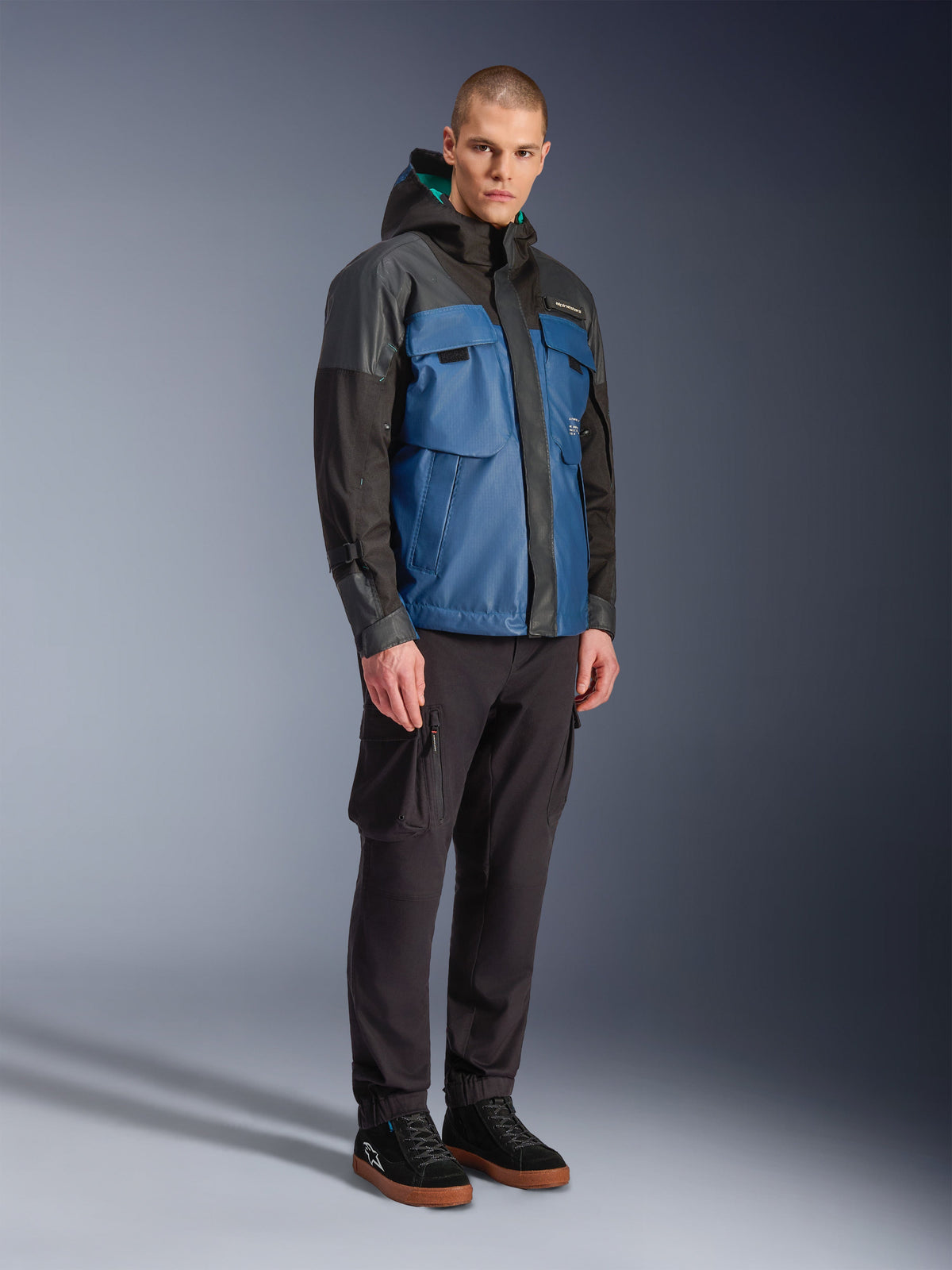 Chaqueta impermeable Alpinestars Mospher, negro y azul petróleo oscuro, combinada con pantalones cargo negros y zapatillas negras, estilo urbano con bolsillos utilitarios y capucha