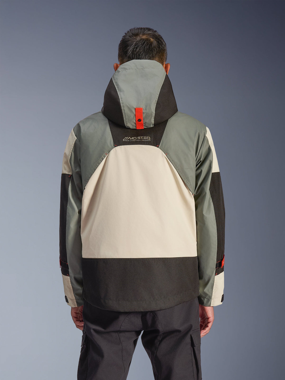 Vista trasera de la chaqueta impermeable Alpinestars Mospher en color Fur Gray Black Ice, con un diseño de paneles en gris, crema y negro, silueta de parka con capucha con detalle de anclaje de botón rojo, marca 'MO.ST.EQ.', combinada con pantalones de conducción tipo cargo negros