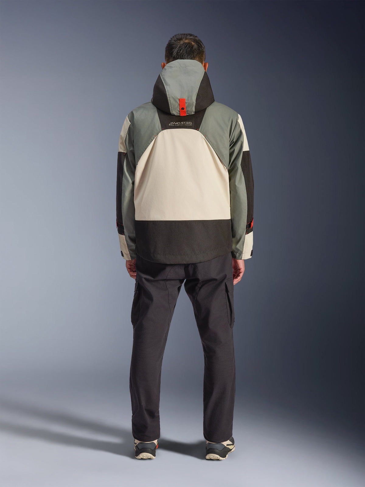 Vista trasera de un piloto con la chaqueta Alpinestars Mospher Waterproof en Fur Gray Black Ice, parka de inspiración táctica con bloques de color gris, negro y crema, incluye una capucha con detalle de correa naranja, combinada con pantalones cargo negros.