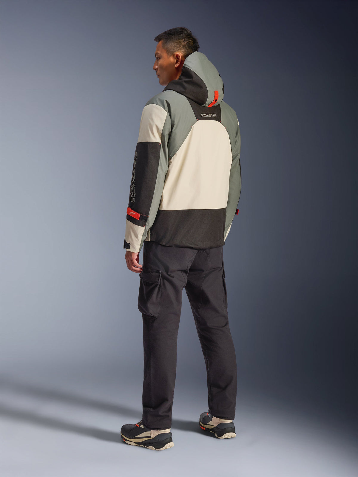 Chaqueta Alpinestars Mospher Waterproof en color Fur Gray Black Ice, vista posterior de un modelo con una parka táctica de motociclismo con paneles en crema, gris y negro, detalles rojos y capucha, combinada con pantalones cargo negros y botas técnicas