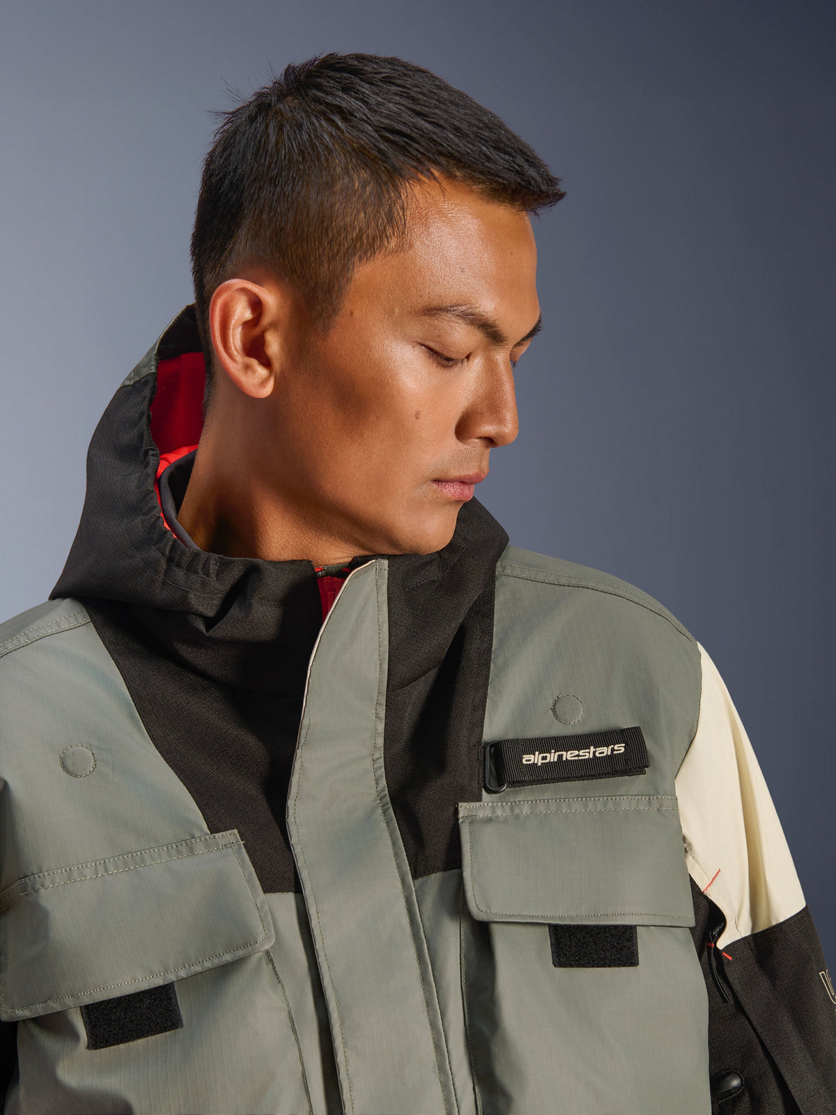 Chaqueta Alpinestars Mospher WP, parka de moto impermeable, Fur Gray Black Ice, primer plano de modelo usando chaqueta con capucha y bolsillos utilitarios, etiqueta con logo de Alpinestars en negro y diseño táctico con forro interior rojo