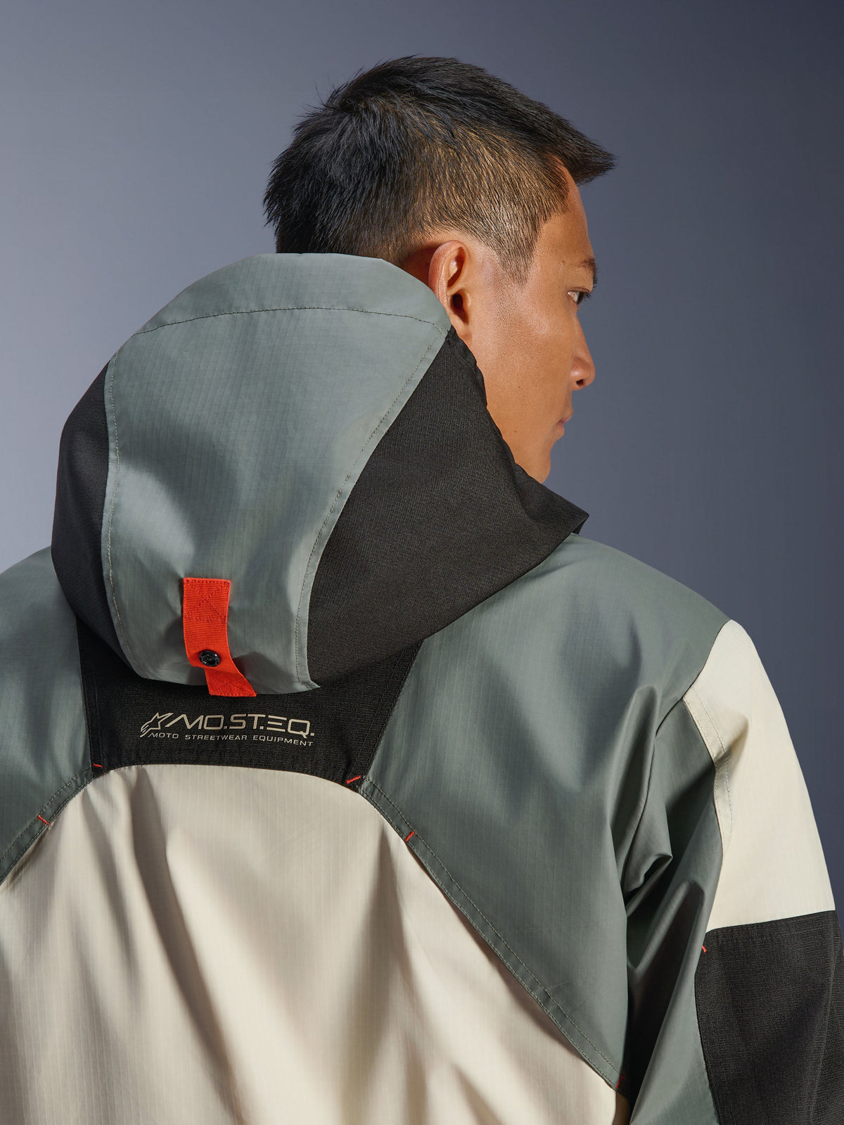 Chaqueta Alpinestars Mospher Waterproof, Fur Gray Black Ice, primer plano de la vista trasera que muestra el sistema de anclaje de la capucha con una lengüeta de botón de presión naranja, marca MO.ST.EQ. Moto Streetwear Equipment, construcción de tejido ripstop duradero