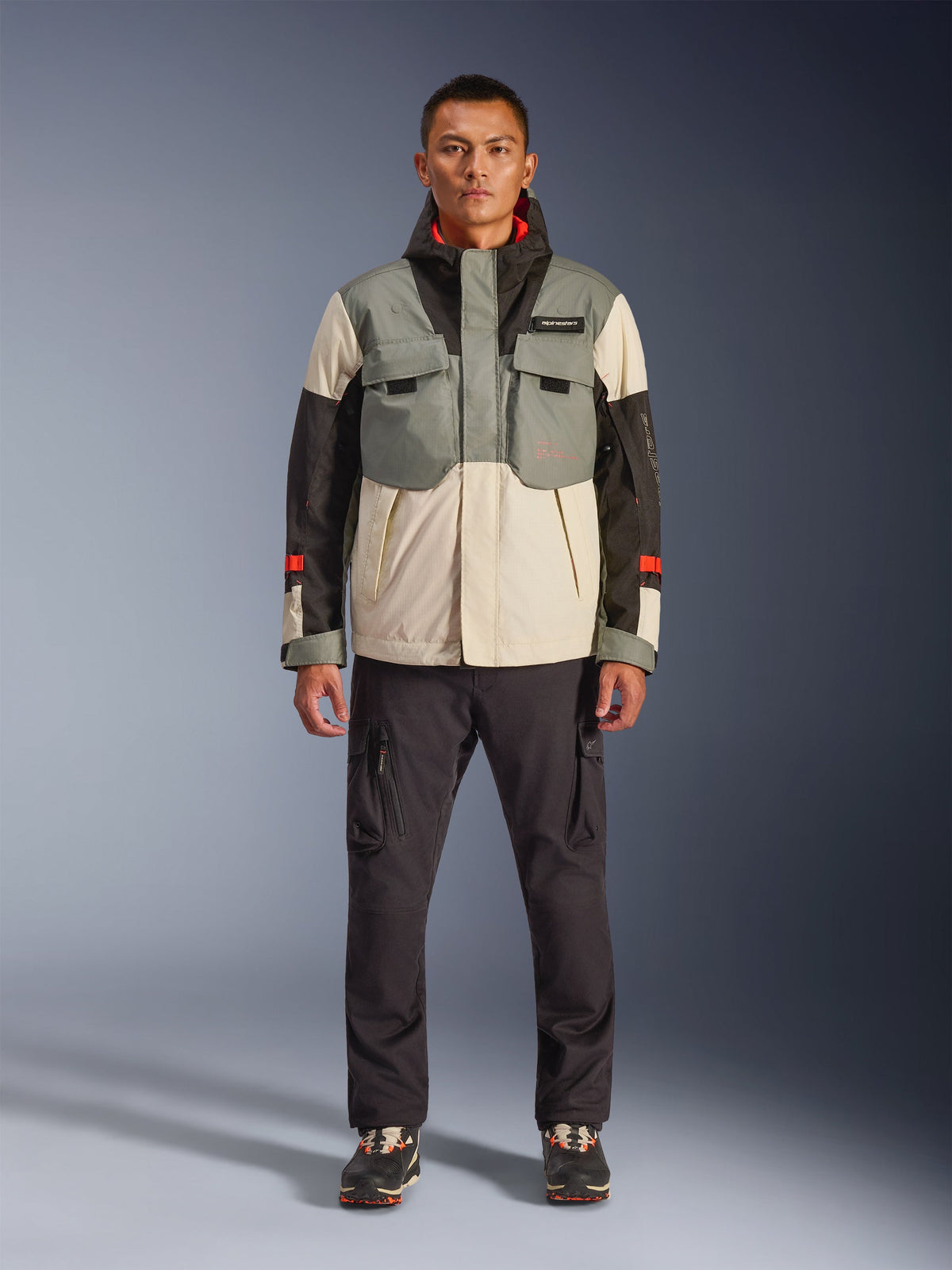 Chaqueta impermeable Alpinestars Mospher en Fur Gray Black Ice, parka de moto de estilo táctico usada por un modelo masculino con pantalones cargo oscuros, diseño con múltiples bolsillos y paneles en gris, crema y negro, equipamiento de conducción urbana