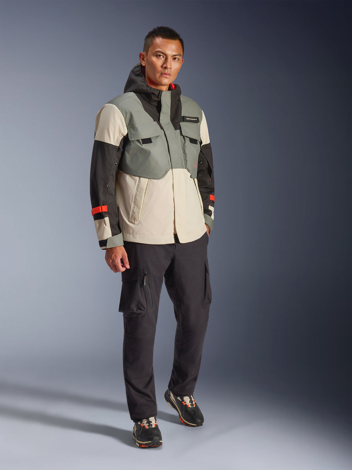 Alpinestars Mospher Waterproof Jacket en Fur Gray Black Ice, parka técnica de motociclismo multicolor con paneles grises, negros y blanco roto y acentos rojos, diseño funcional con grandes bolsillos y capucha, combinada con pantalones cargo gris oscuro y zapatillas negras, equipamiento de motociclismo urbano contemporáneo