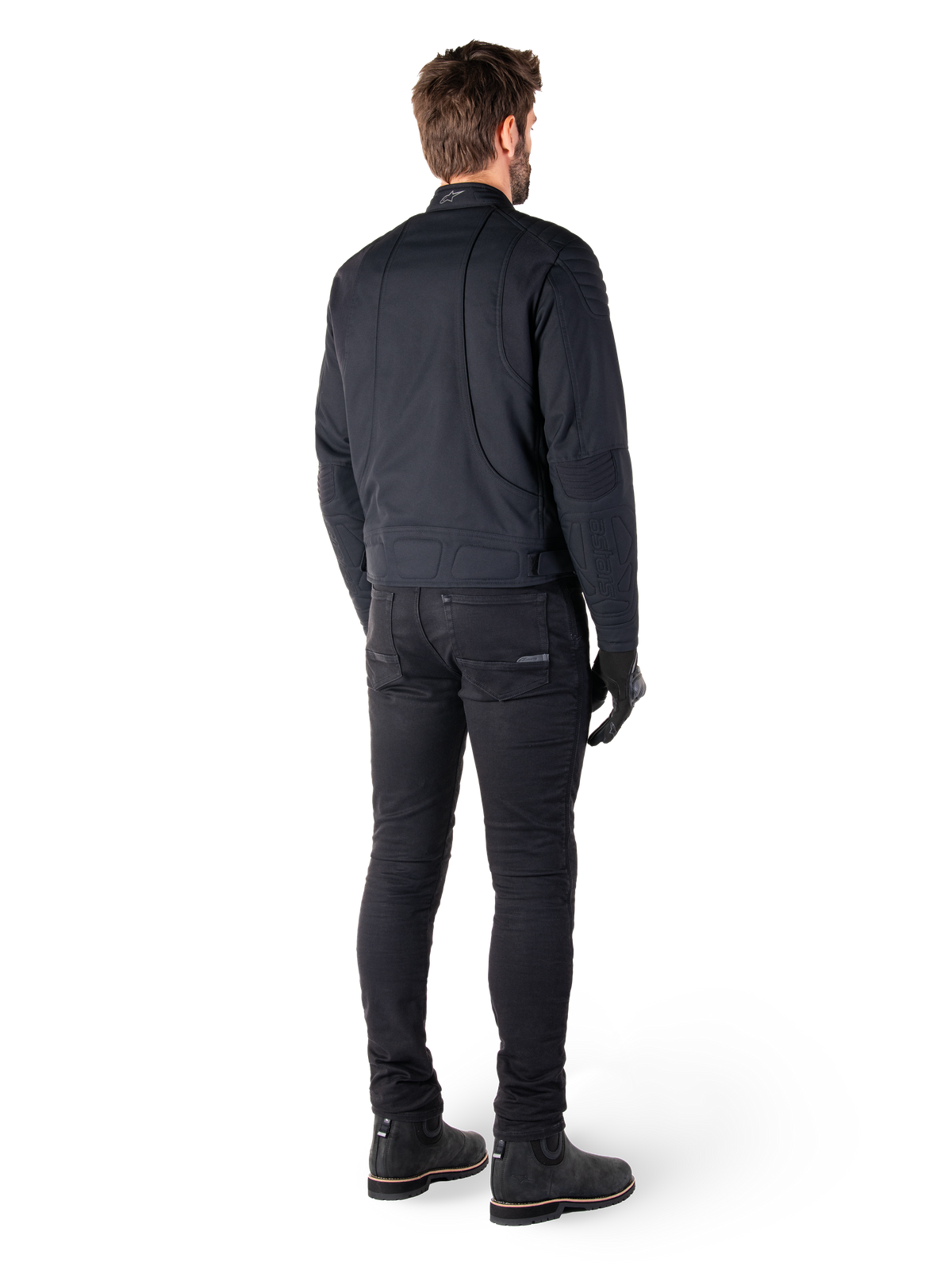 Vista posterior de un motorista con la chaqueta Alpinestars Clayton WR en negro, paneles acolchados en codos y hombros, costuras ergonómicas curvas y detalles reflectantes en el cuello, combinada con pantalones y botas negras para un look urbano de motociclista