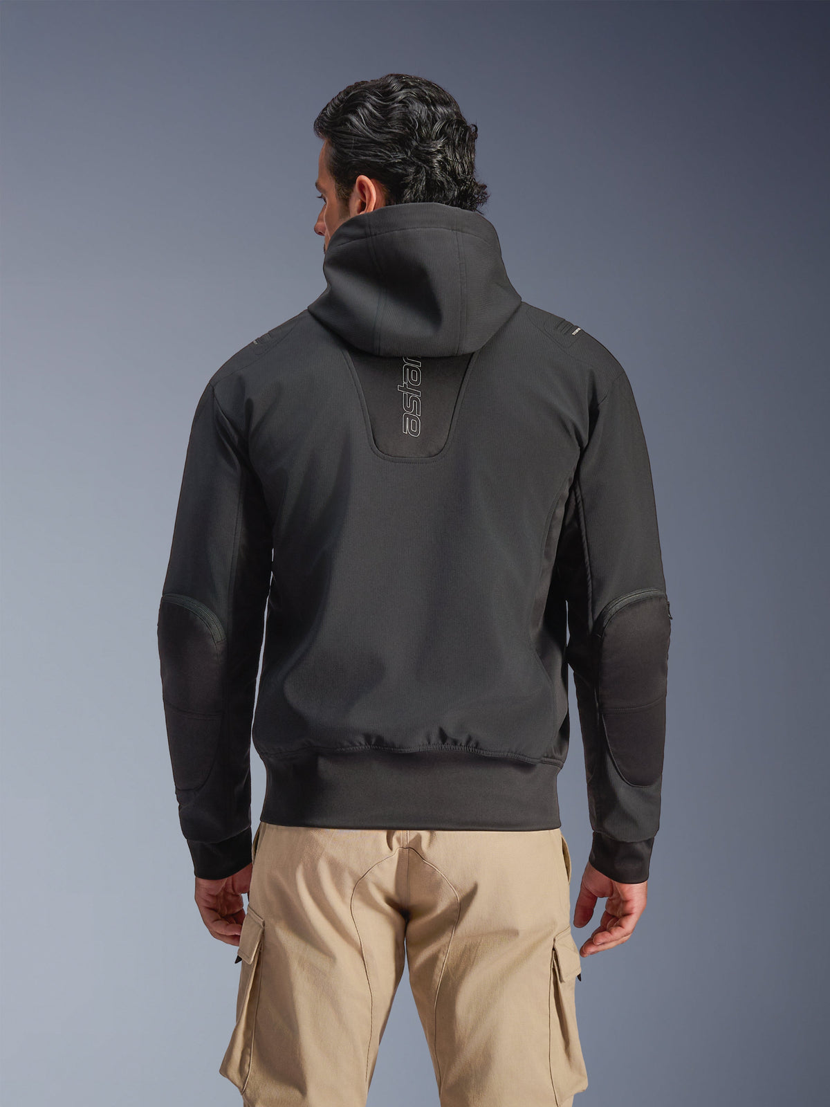 Chaqueta impermeable Alpinestars Moblast, negra, vista trasera de chaqueta de moto urbana con capucha, con logotipo vertical 'astars' en la espalda, cintura y puños elásticos, codos reforzados y diseño tecnológico moderno, usada con pantalones cargo color canela.