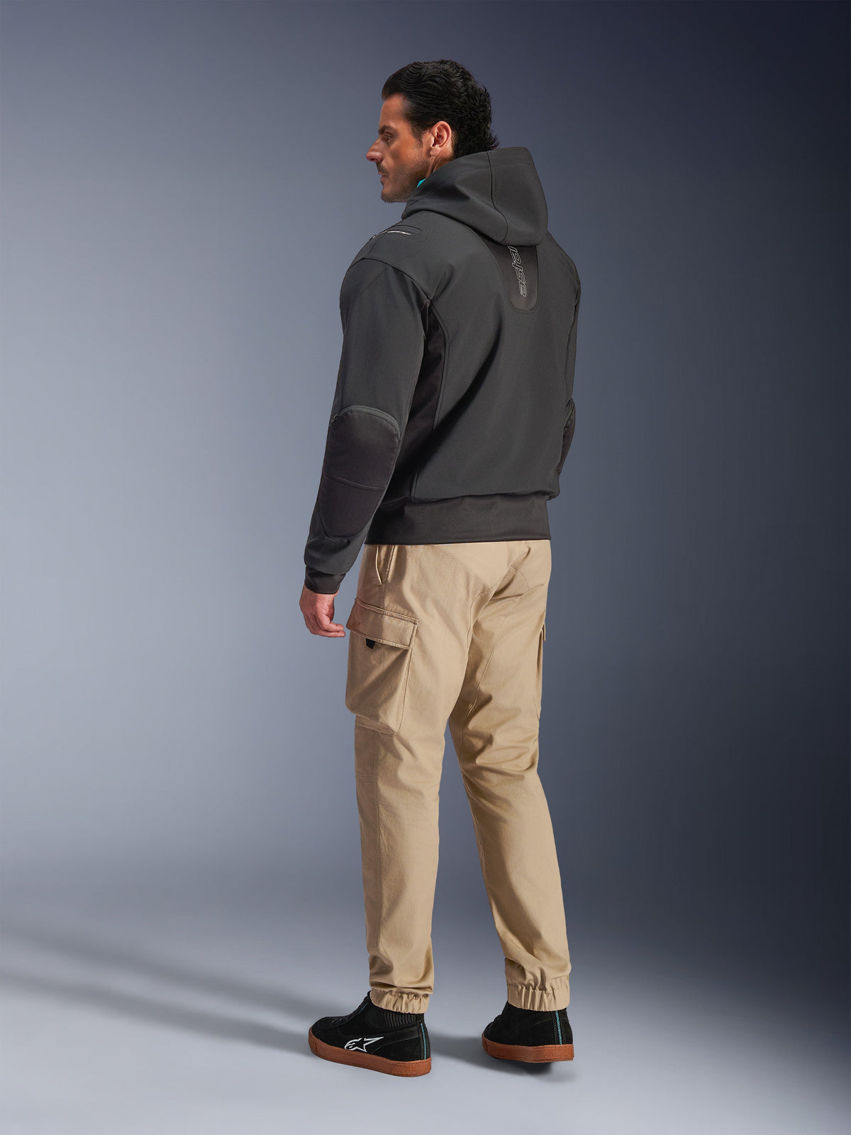 Chaqueta Alpinestars Moblast Waterproof, sudadera técnica de moto negra, vista lateral y trasera, combinada con pantalones cargo color canela y zapatillas negras, con un diseño con capucha, construcción textil técnica y una estética urbana moderna.
