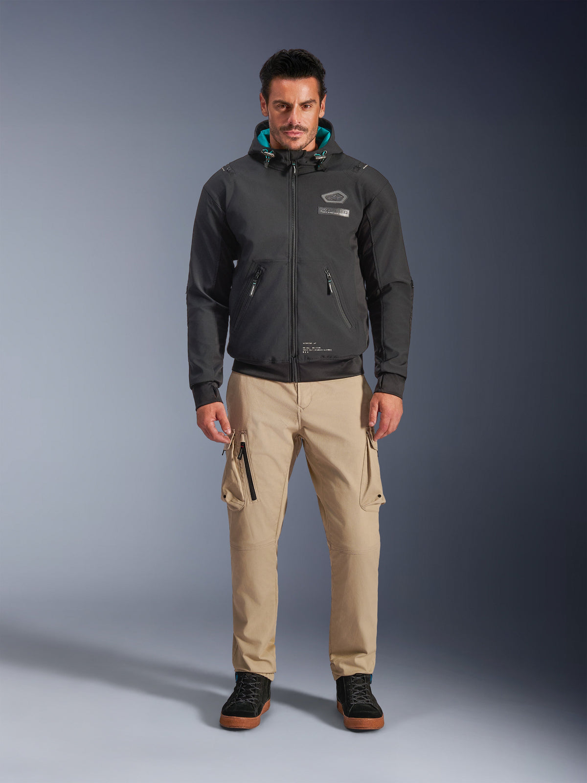 Chaqueta impermeable Alpinestars Moblast, sudadera de moto urbana negra con forro de la capucha en color verde azulado, usada con pantalones cargo color canela y botas altas negras, equipamiento de conducción contemporáneo para todas las estaciones