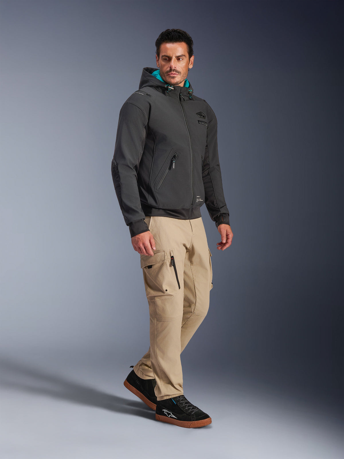 Modelo masculino con la chaqueta Alpinestars Moblast Waterproof Jacket en color negro, sudadera urbana para moto con capucha forrada en verde azulado, combinada con pantalones cargo caqui y zapatillas negras, diseño minimalista moderno con tejido técnico elástico para desplazamientos en cualquier clima