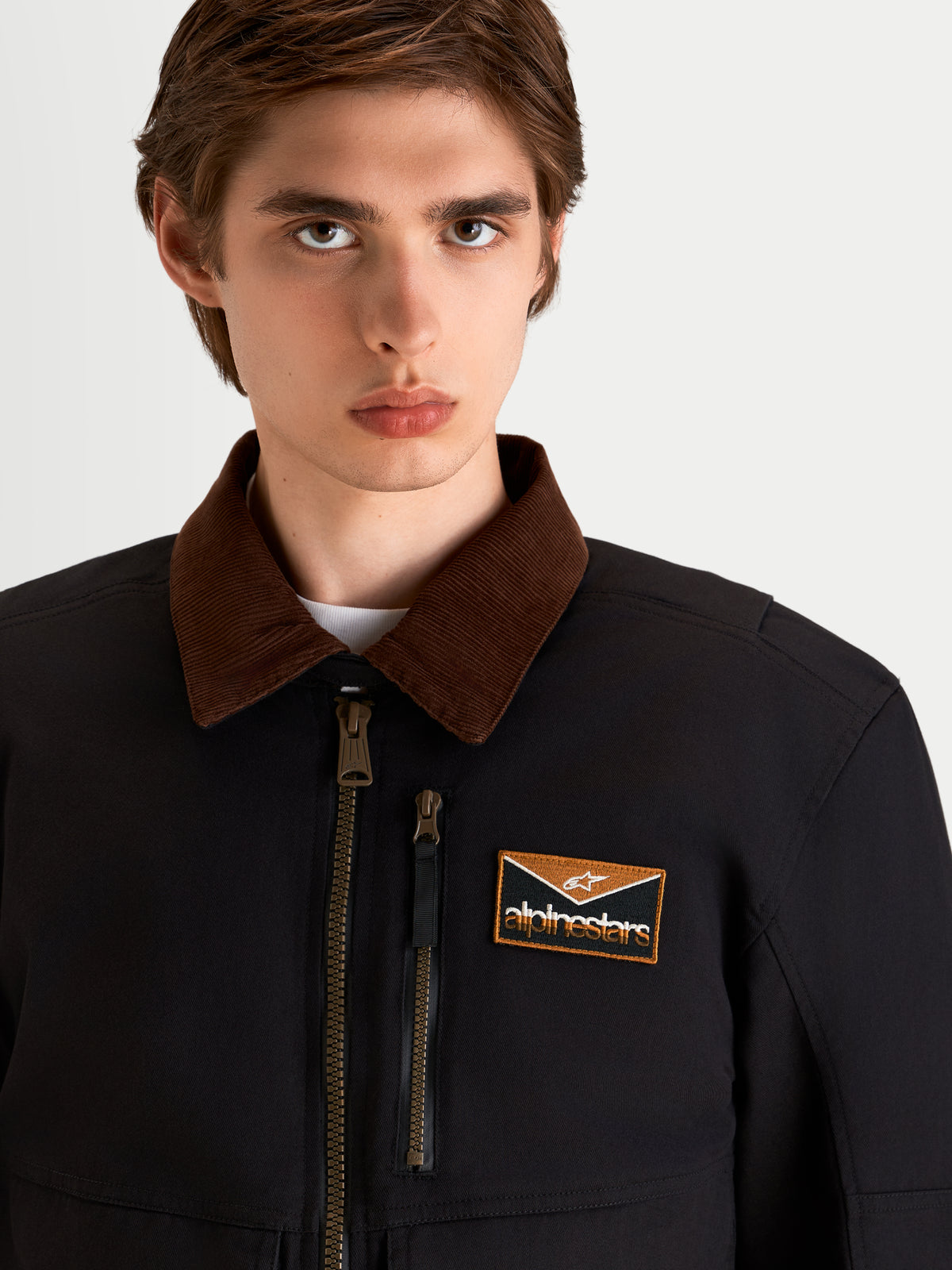 Alpinestars Mustang Chaqueta de moto impermeable para hombre, en antracita oscuro y marrón oscuro, primer plano con cuello de pana de inspiración workwear, cremallera frontal y parche con el logo en el pecho en un modelo