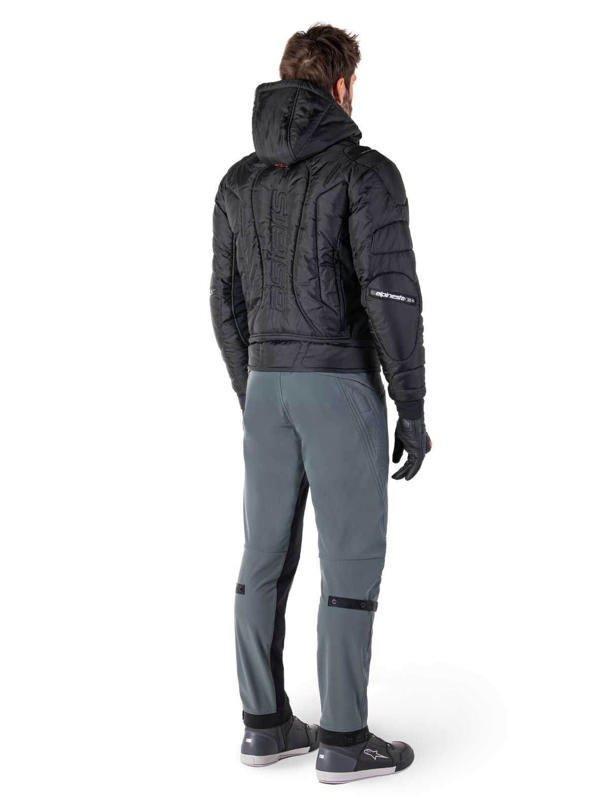 Chaqueta impermeable Alpinestars Mohobbs, negra, vista trasera de chaqueta bomber acolchada con logotipo vertical en relieve en la espalda, combinada con pantalones de conducción grises y guantes negros, equipamiento de motociclismo de estilo urbano
