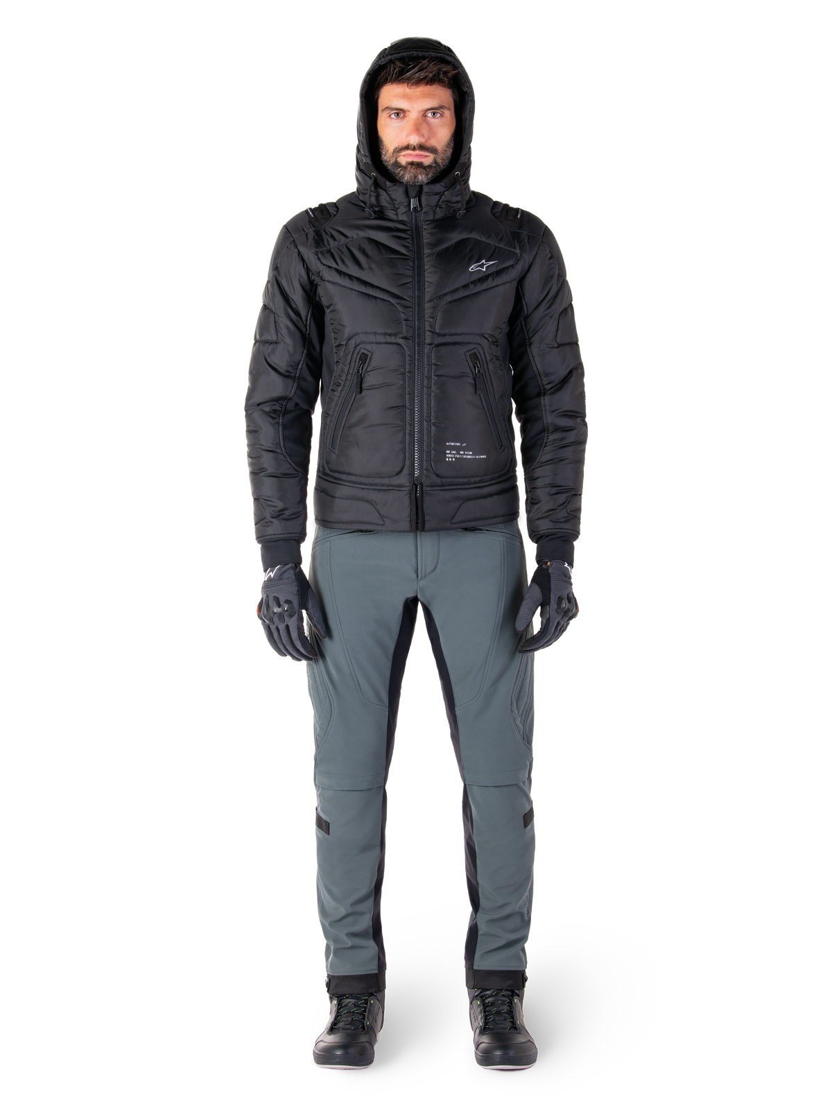 Chaqueta Alpinestars Mohobbs Waterproof en color negro, modelo masculino con chaqueta bomber acolchada y capucha, combinada con pantalones de conducción grises, guantes negros y botas negras, equipamiento de motociclismo urbano contemporáneo.