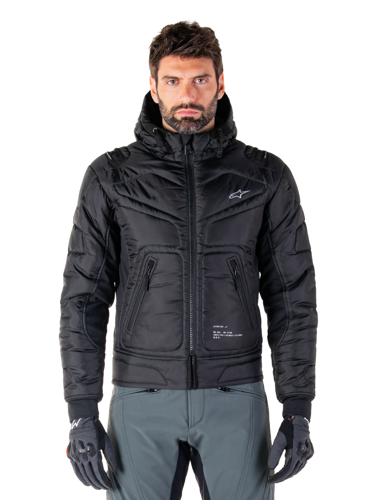 Alpinestars Mohobbs Waterproof Jacket, chaqueta bomber de moto negra acolchada, tejido de poliéster acolchado con cremallera frontal y bolsillos con cremallera, pequeño logo blanco en el pecho, usada por un modelo con pantalones grises y guantes, diseño de estilo urbano para conducción en ciudad