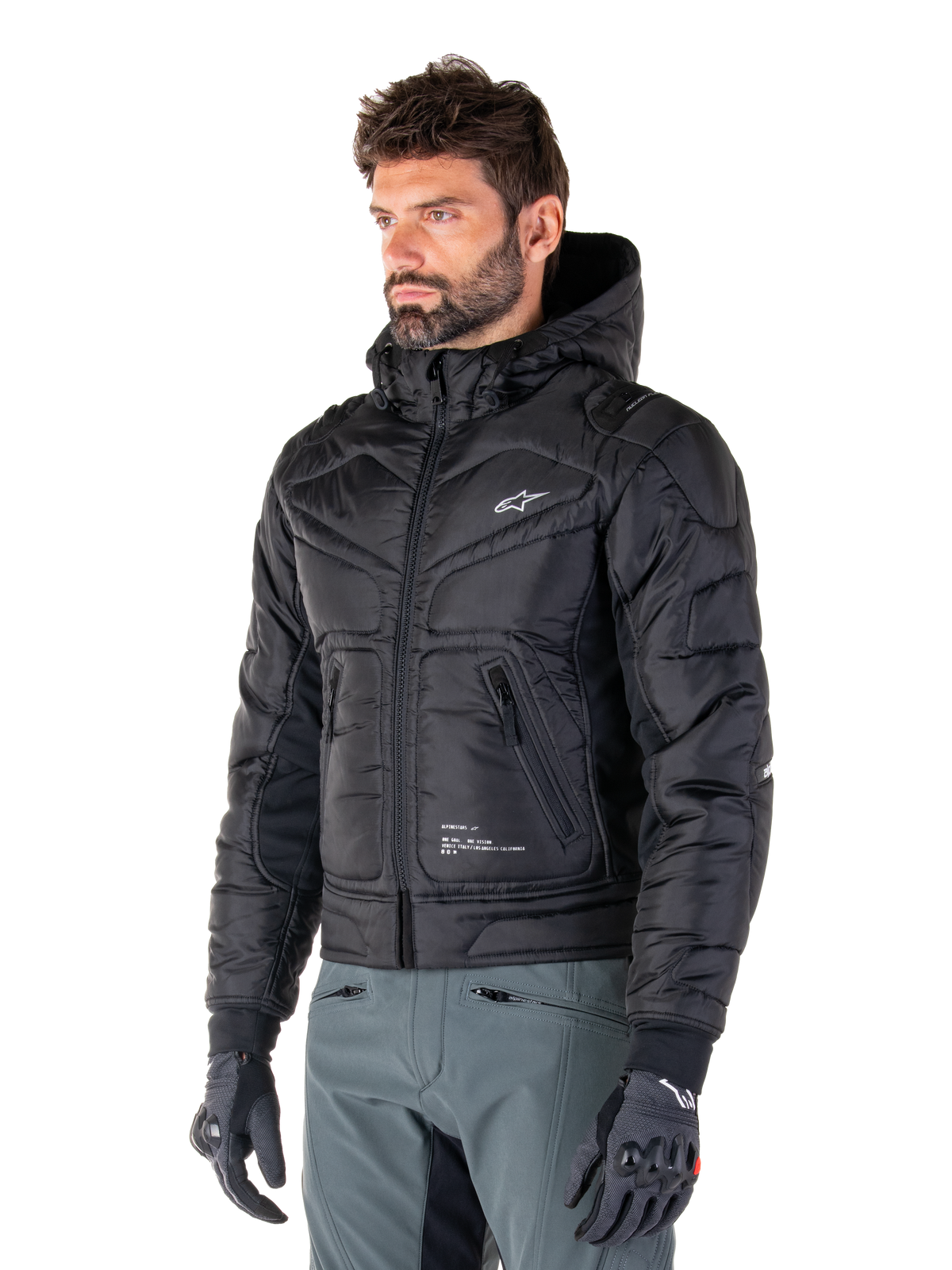 Chaqueta impermeable Mohobbs de Alpinestars, negra, diseño de chaqueta bomber acolchada con capucha, usada por un hombre con pantalones de moto grises y guantes negros, ropa de moto de inspiración urbana con un pequeño logo blanco en el pecho