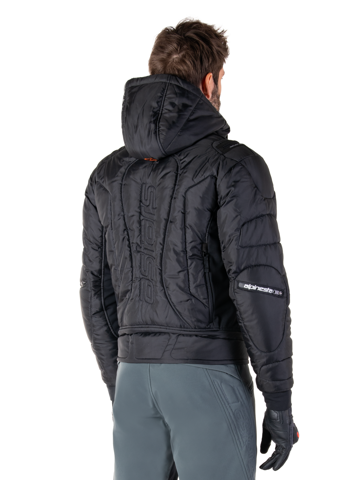 Chaqueta Alpinestars Mohobbs Waterproof, negra, vista trasera que muestra la construcción acolchada, capucha, marca denominativa 'astars' vertical en la espalda y logotipos de Alpinestars en las mangas, usada con pantalones grises y guantes negros.