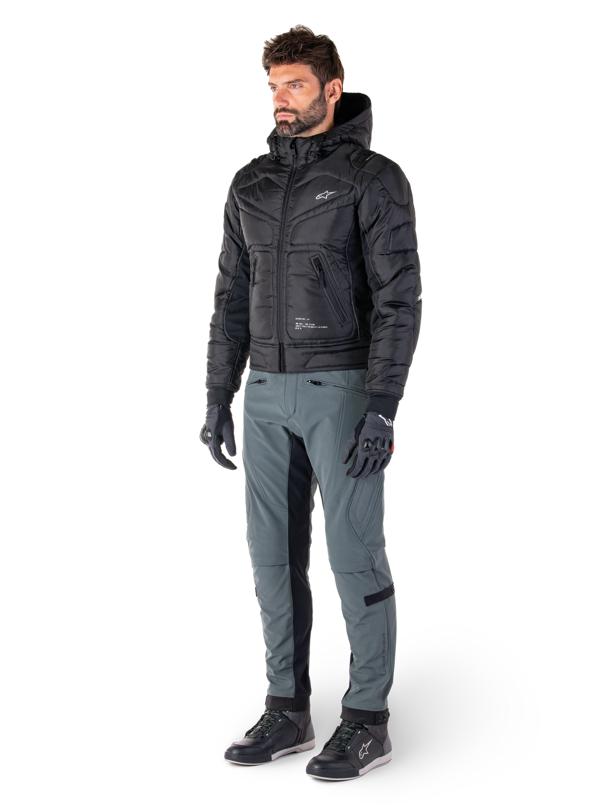 Chaqueta impermeable Alpinestars Mohobbs, negra, chaqueta bomber de moto urbana acolchada con capucha, usada por un modelo con pantalones de moto grises y guantes negros, con un logotipo blanco en el pecho, equipo de protección contemporáneo inspirado en el estilo streetwear