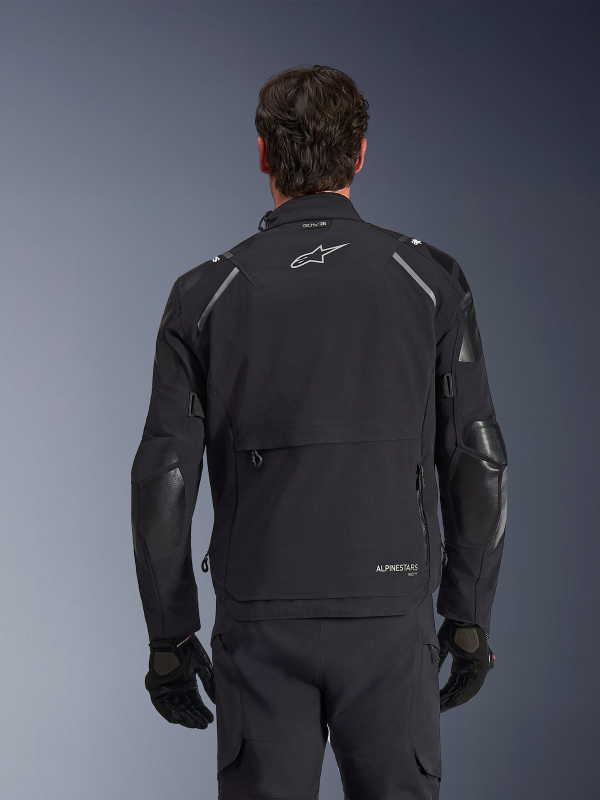 Alpinestars Andes Pro Drystar® XF Jacket, Black and Dark Gray, back view worn by a male model, featuring a large rear storage compartment, reflective shoulder accents, and logo branding, paired with black gloves and pants for adventure touring.