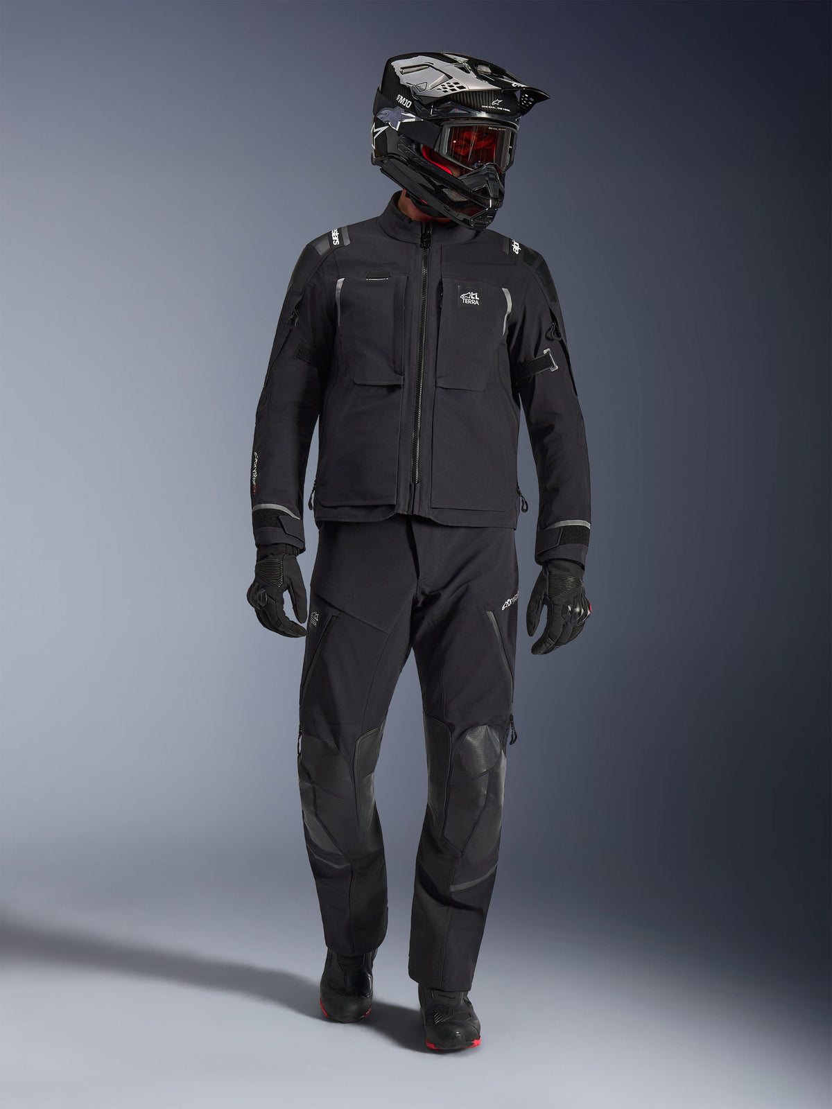 Adventure rider wearing the Alpinestars Andes Pro Drystar® XF Jacket in Black and Dark Gray, matching black adventure pants, black and white helmet with red goggles, black gloves, and boots, features large front utility pockets, reflective accents, and technical 3-layer laminated construction for all-weather touring.