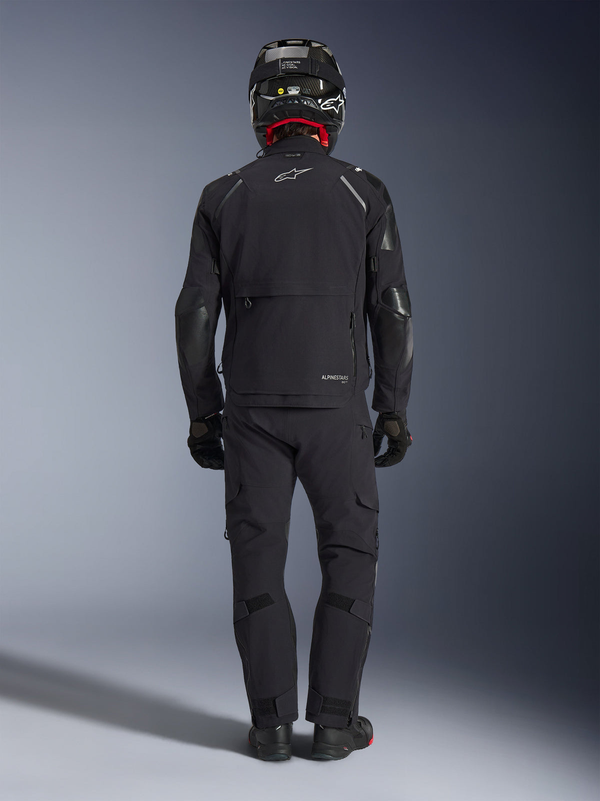 Alpinestars Andes Pro Drystar® XF Jacket in Black and Dark Gray, back view of a rider in full adventure touring gear including matching pants and carbon fiber helmet, featuring a large rear cargo pocket and reflective Alpinestars branding on the back.