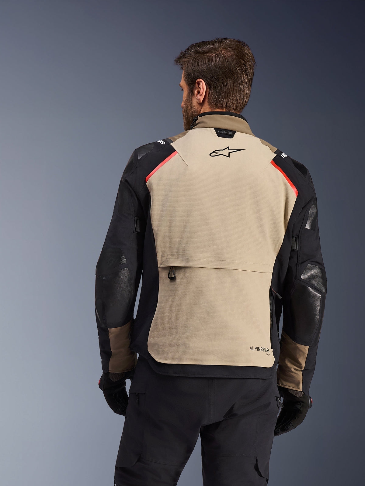 Chaqueta Alpinestars Andes Pro Drystar® XF, vista trasera, color Walnut y Aluminum, con un gran panel trasero en color canela, logotipo de Alpinestars en negro y detalles rojos en los hombros, gran bolsillo de carga trasero, combinada con pantalones y guantes negros, equipamiento de motociclismo para turismo de aventura