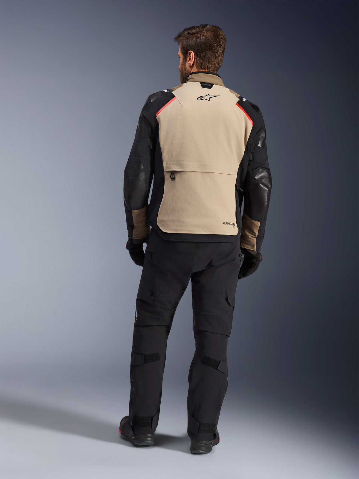 Chaqueta Alpinestars Andes Pro Drystar® XF, Walnut Aluminum, vista trasera, chaqueta de aventura para motocicleta con mangas negras y detalles rojos en los hombros, usada con pantalones de conducción y guantes negros