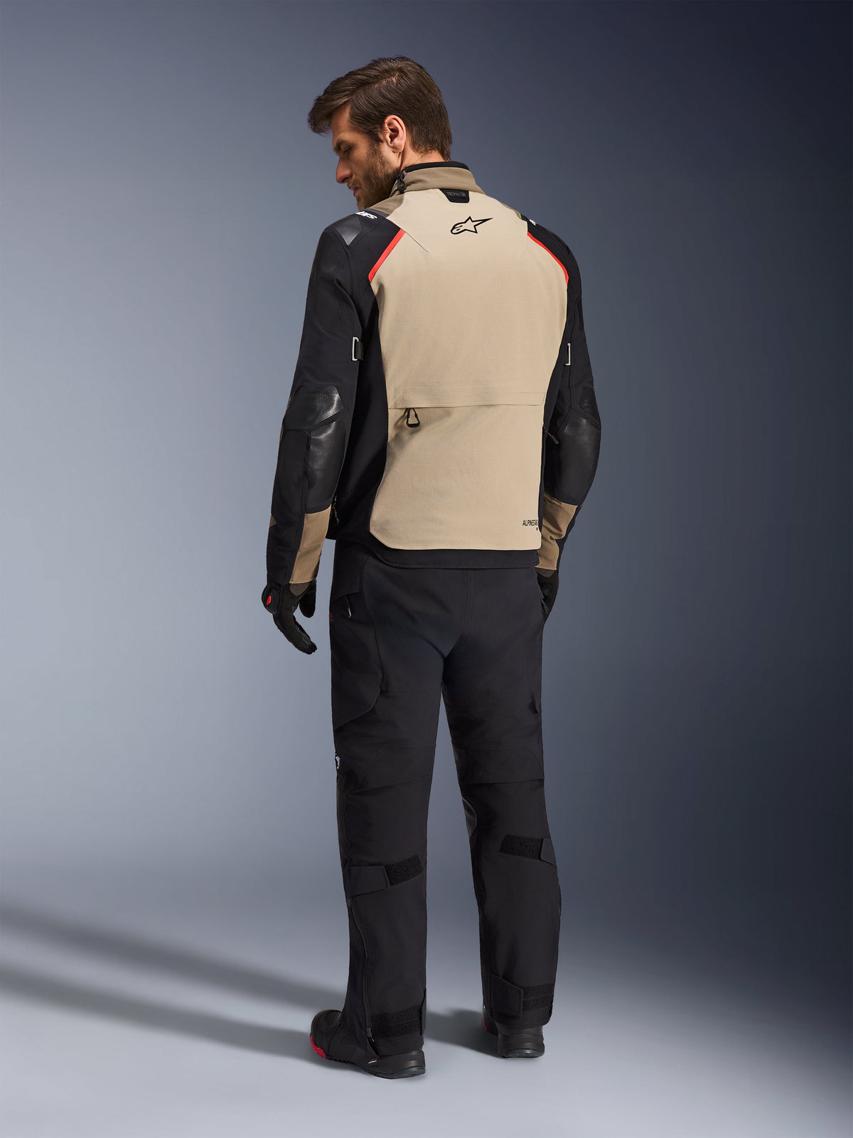 Chaqueta Alpinestars Andes Pro Drystar® XF, chaqueta de motociclismo, color nogal y negro con detalles en rojo, vista trasera en modelo, diseño de turismo de aventura con un gran bolsillo trasero, usada con pantalones de motociclismo y guantes negros.