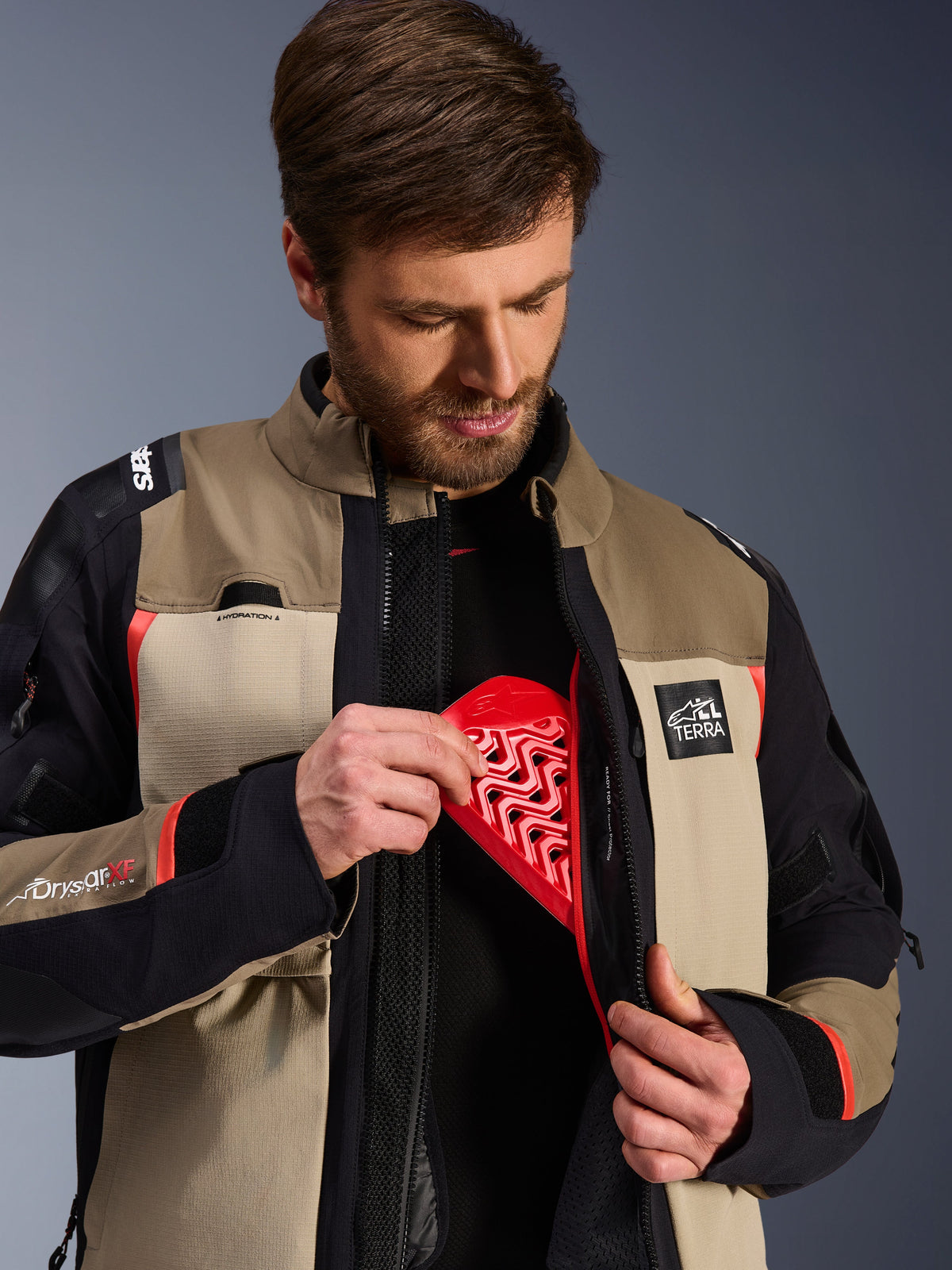 Chaqueta Alpinestars Andes Pro Drystar® XF en Walnut Aluminum, modelo demostrando la inserción del protector de pecho Nucleon rojo en el bolsillo interno, chaqueta de motociclismo de aventura con marca ALL TERRA y logotipos Drystar XF.