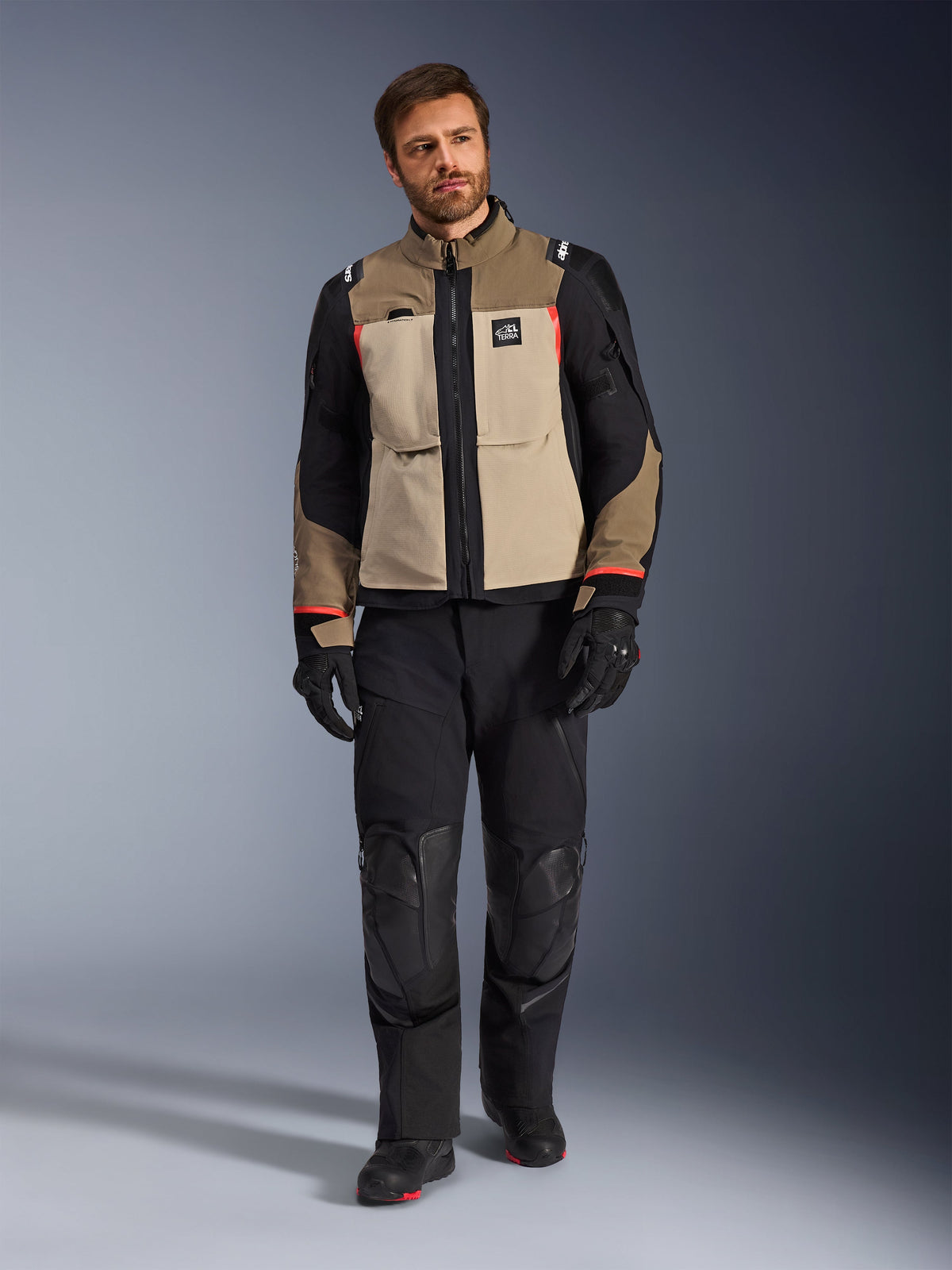 Modelo masculino vistiendo la chaqueta Alpinestars Andes Pro Drystar® XF en Walnut Aluminum, chaqueta de moto adventure touring en color canela y negro con detalles rojos, combinada con pantalones de motociclista, guantes y botas negros, toma de estudio de cuerpo completo de equipamiento de protección.